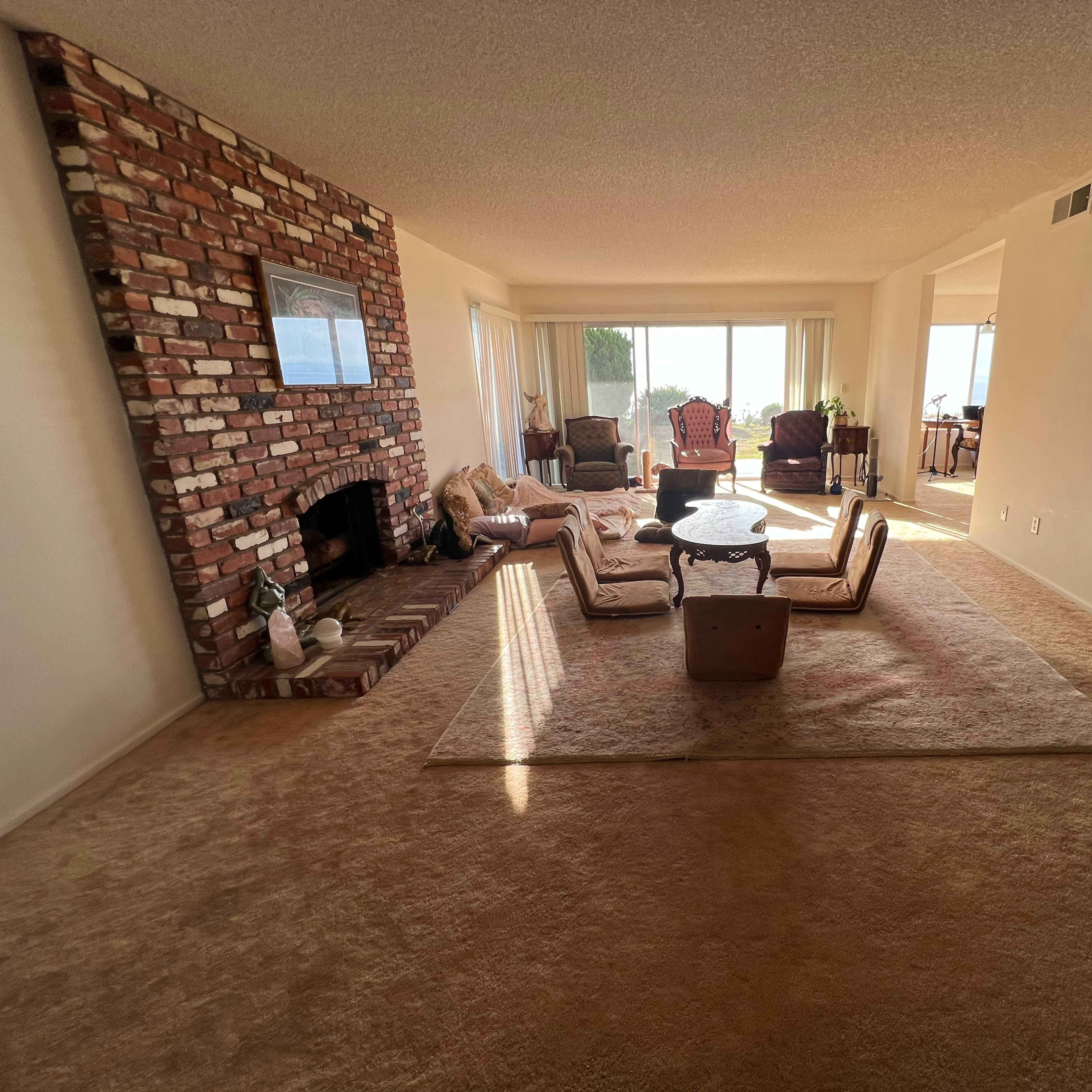 A spacious living room featuring a brick fireplace, a light-colored carpet, and a seating arrangement with a table in the center, illuminated by natural light from large windows.