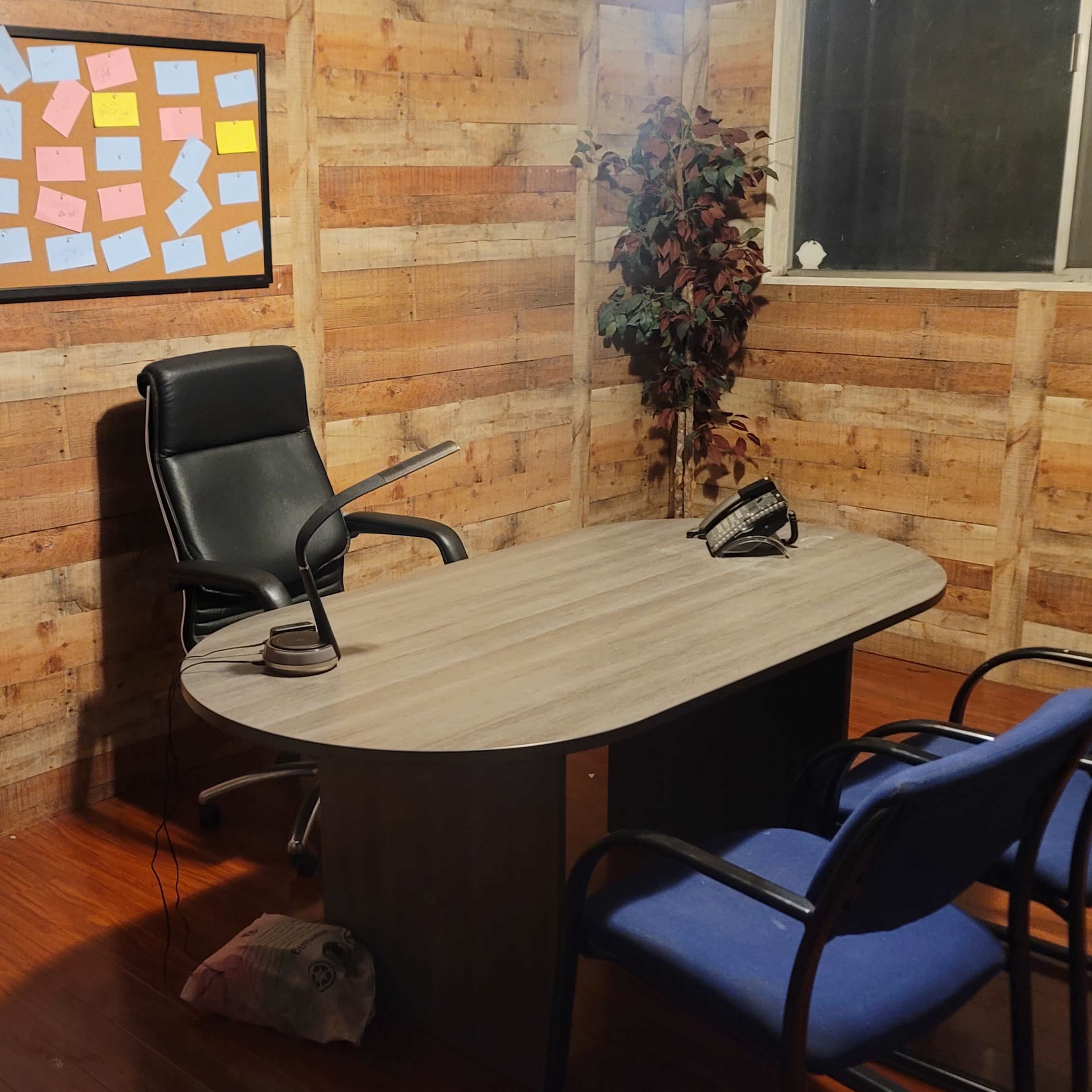 A small office with a round desk, a black chair, two blue chairs, a telephone, a framed board with sticky notes on the wall, and a globe in the corner.