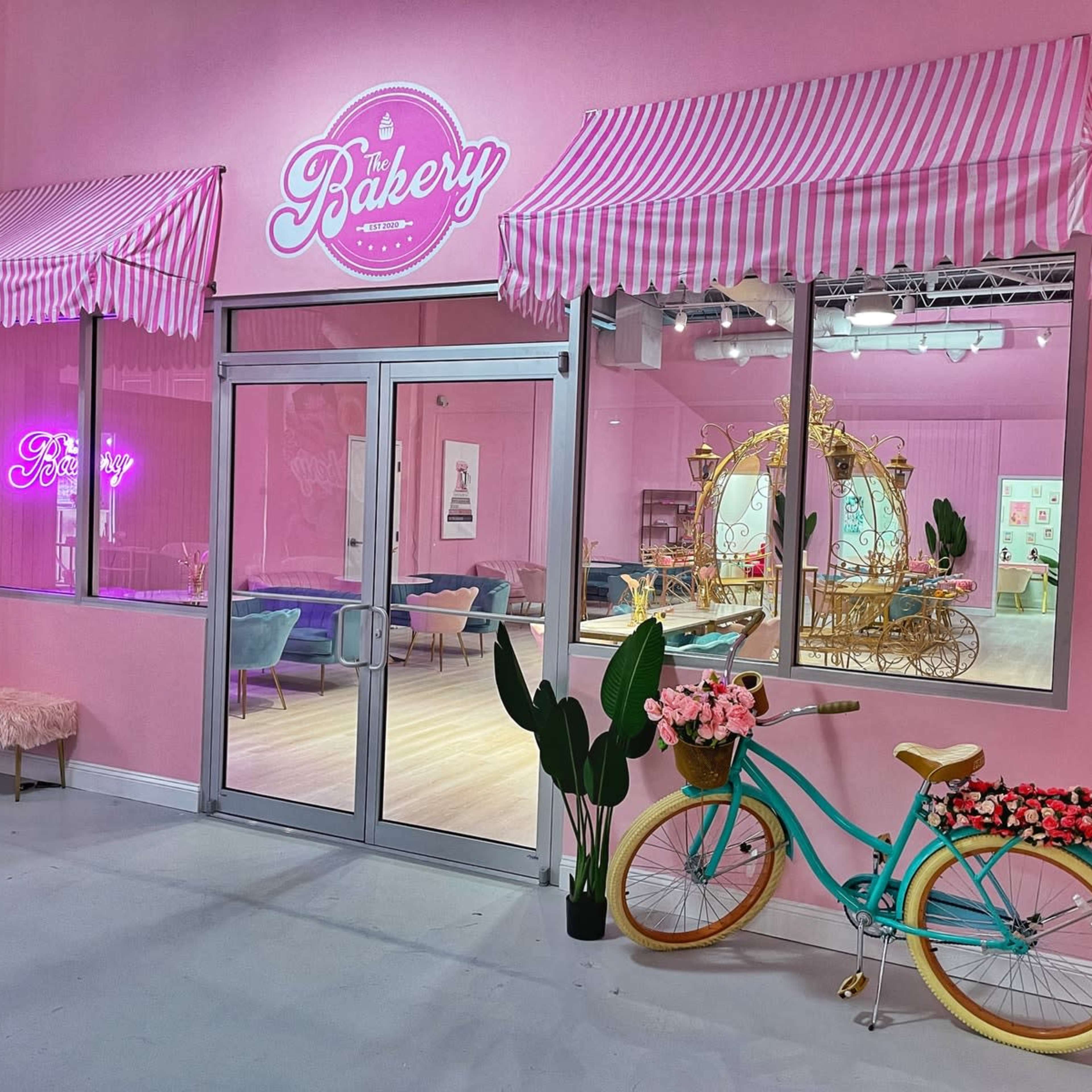A pastel pink bakery storefront features striped awnings, a neon sign, and a decorative bicycle with flowers parked outside.