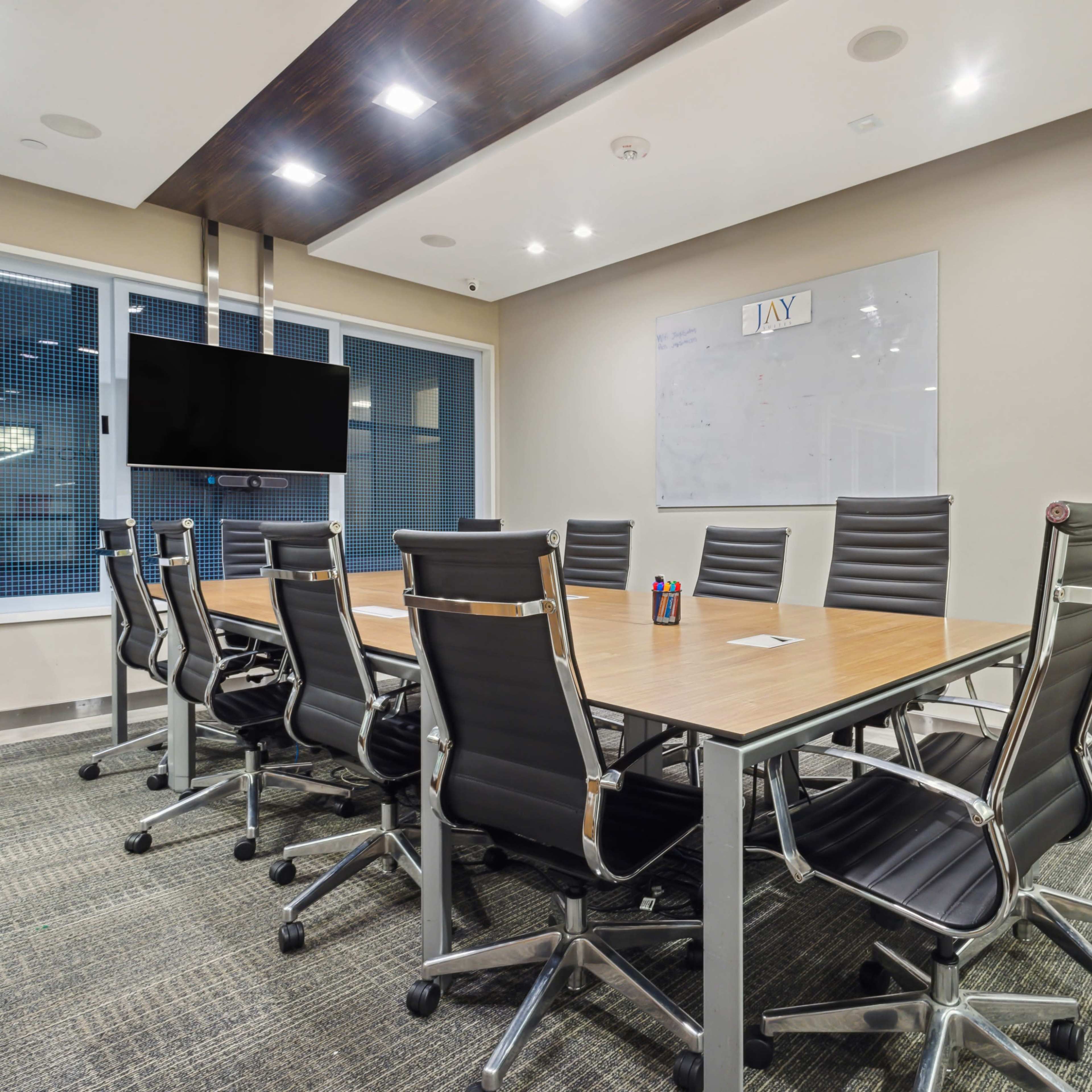 A modern conference room features a long wooden table surrounded by black ergonomic chairs, with a large screen and whiteboard on the wall.
