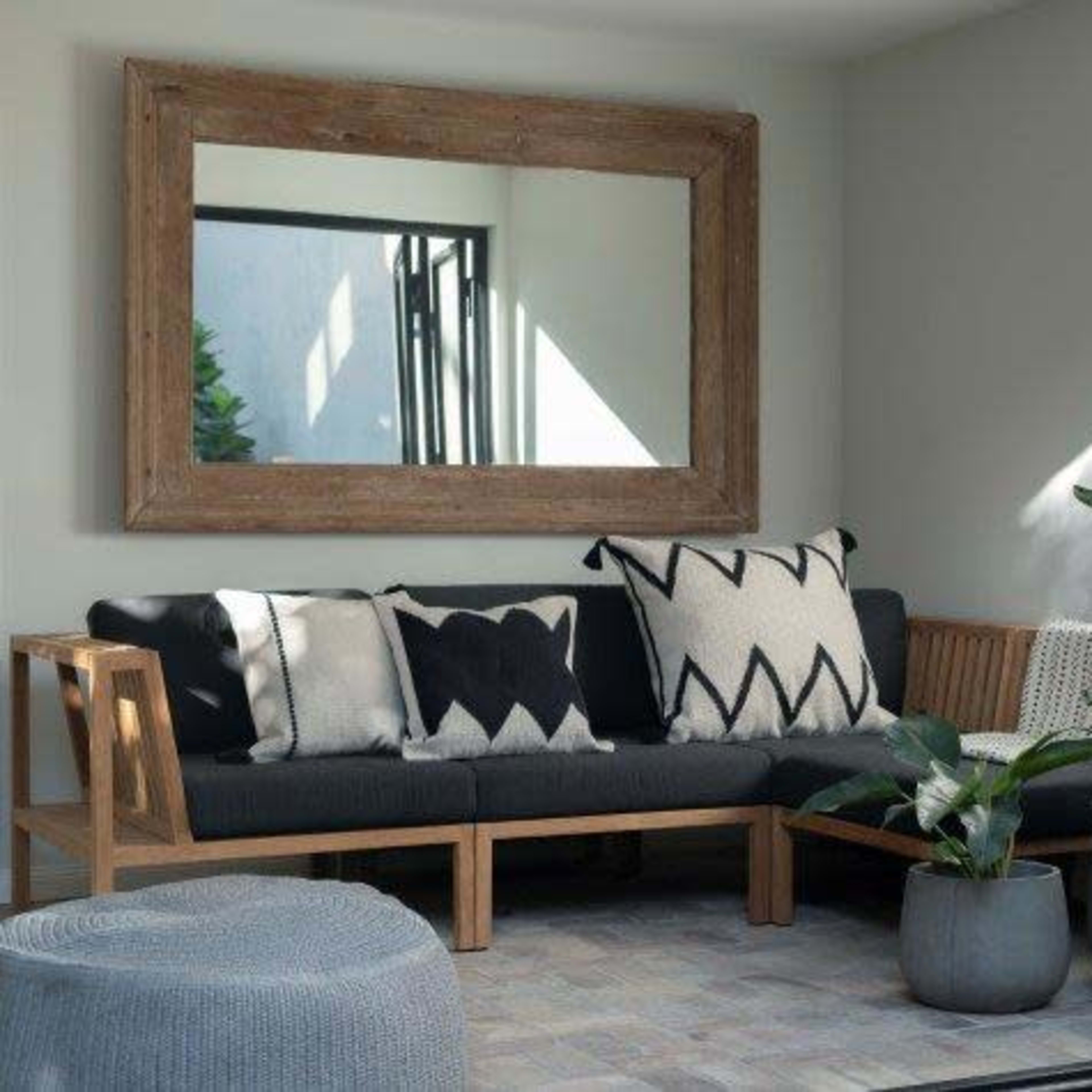A minimalistic living space features a wooden-framed mirror, a sofa with patterned cushions, and potted plants arranged against a light-colored wall.