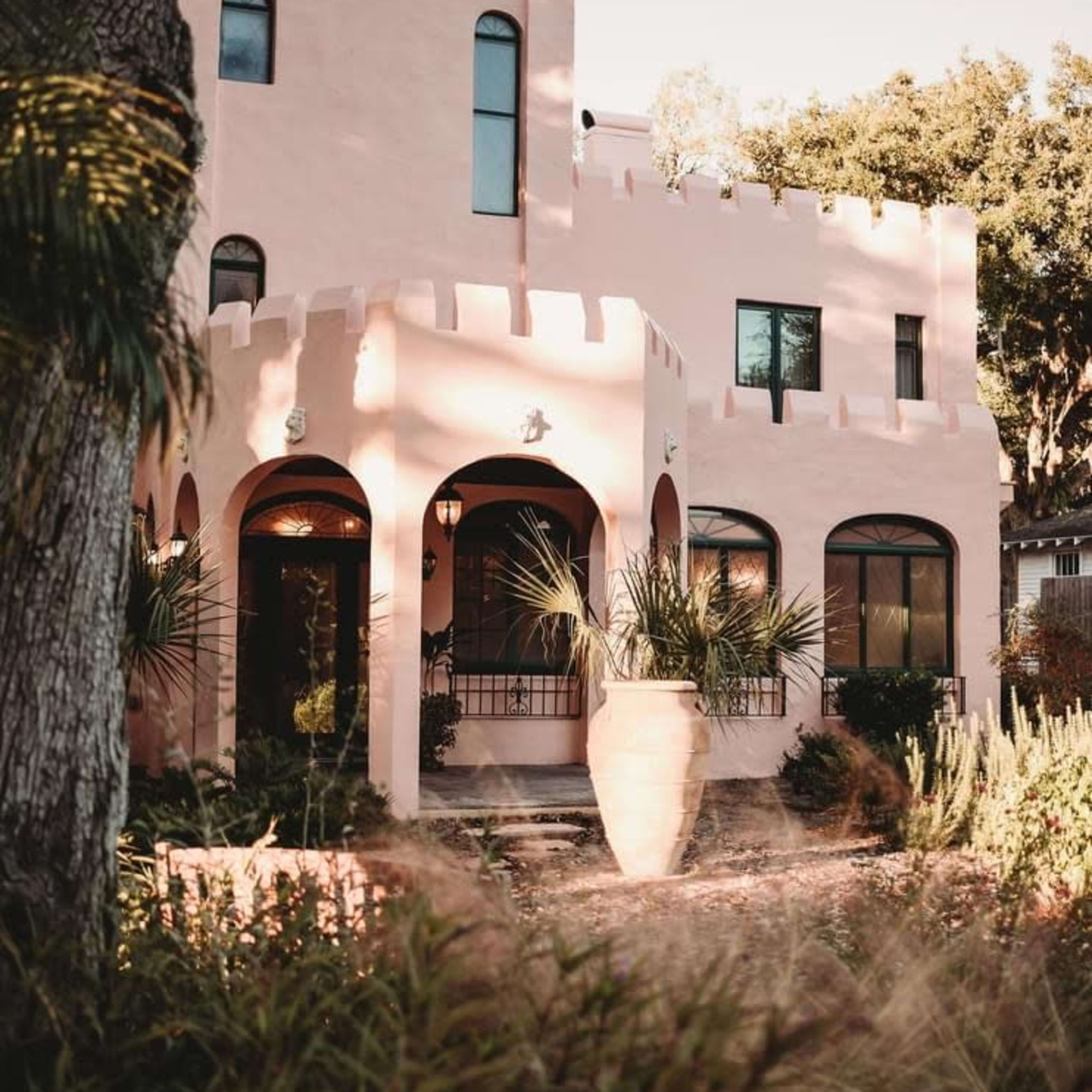 A pink, castle-style house with rounded towers and large arched windows in a lush garden setting.