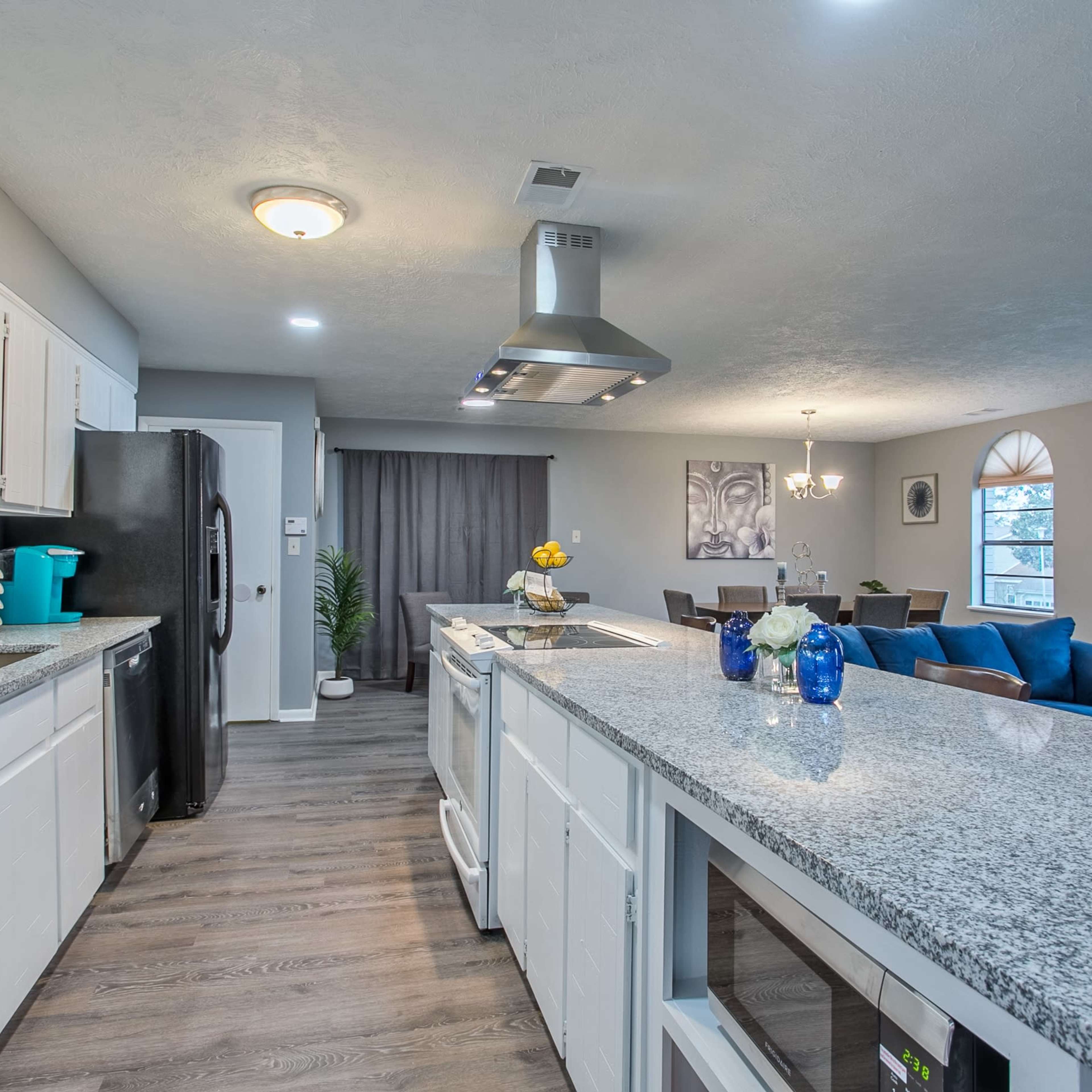 A modern kitchen features a granite countertop with appliances and cabinetry, leading to a dining area with blue sofas and a chandelier.