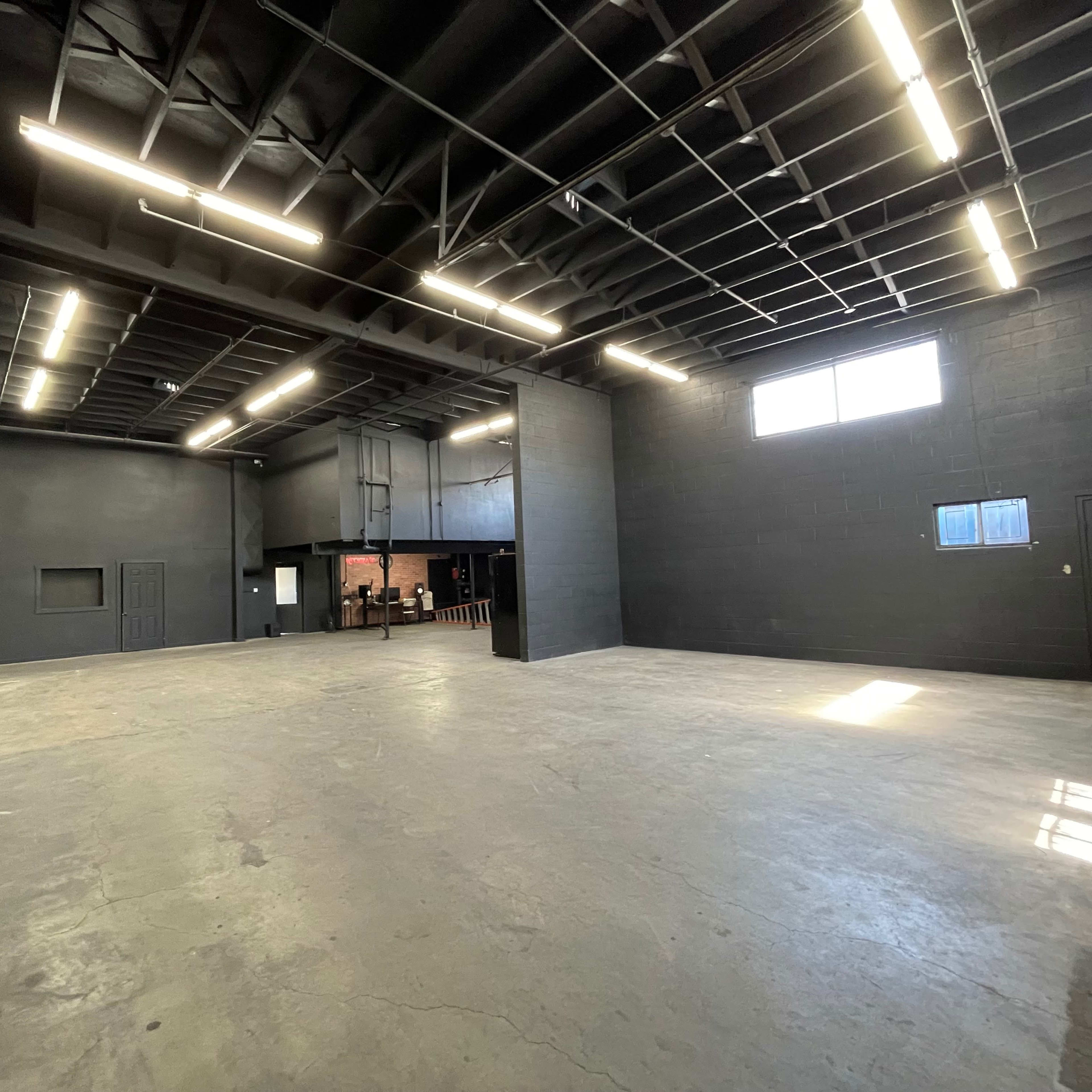 A spacious, empty industrial room with black walls and a concrete floor, featuring overhead lighting and large windows.