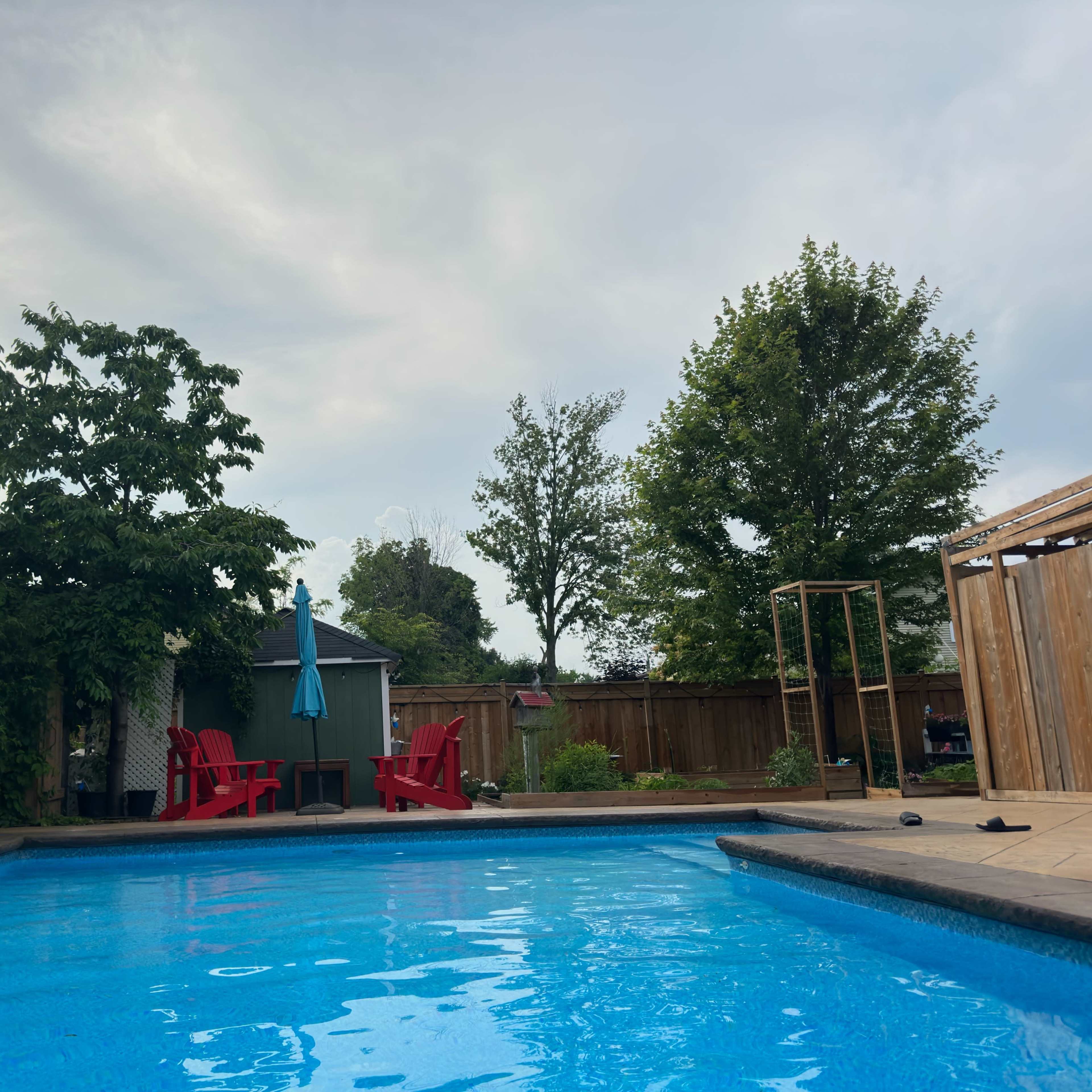A swimming pool with a wooden deck surrounded by greenery and outdoor seating, including red chairs and an umbrella.