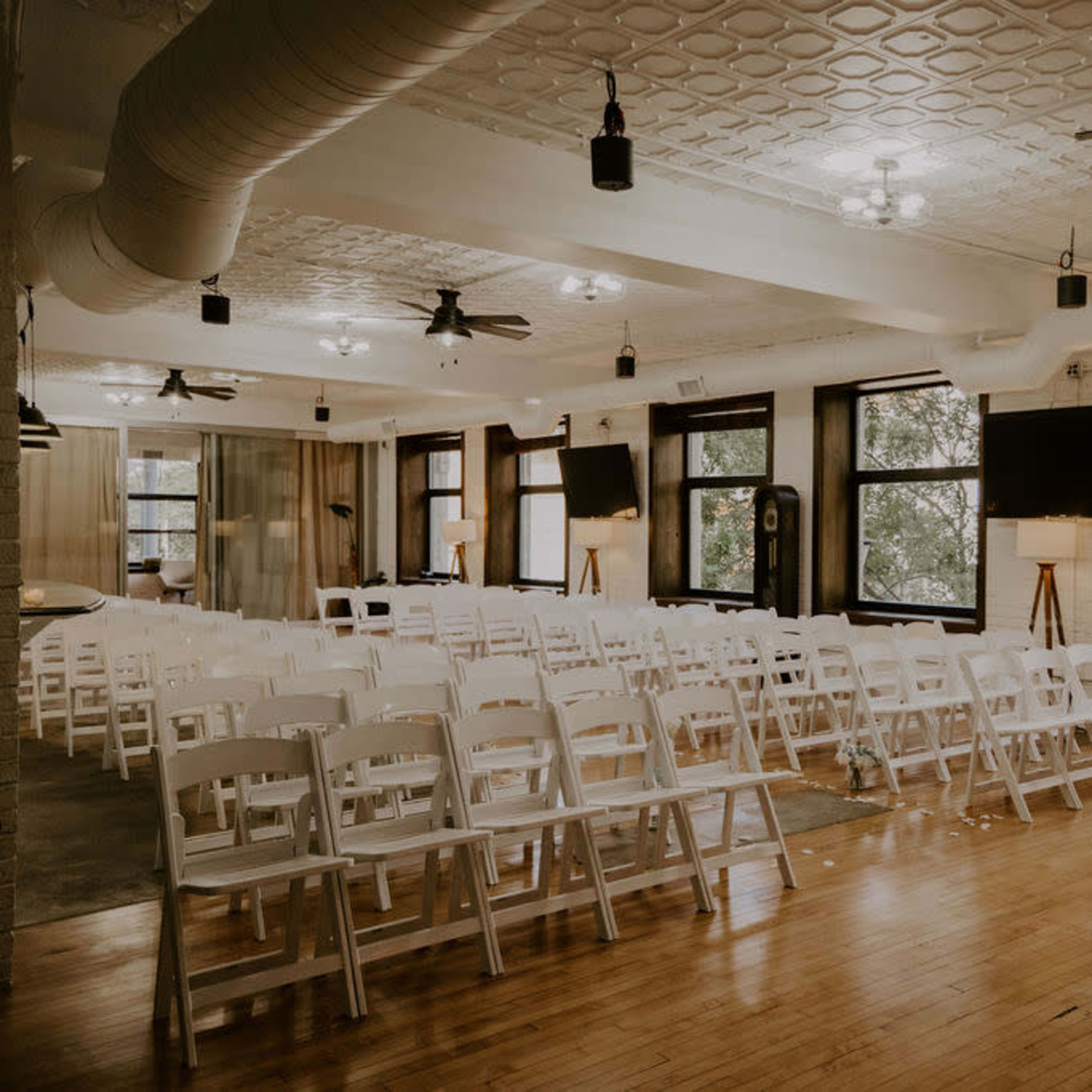 A spacious indoor venue set up with rows of white chairs facing a presentation area, featuring large windows and modern light fixtures.