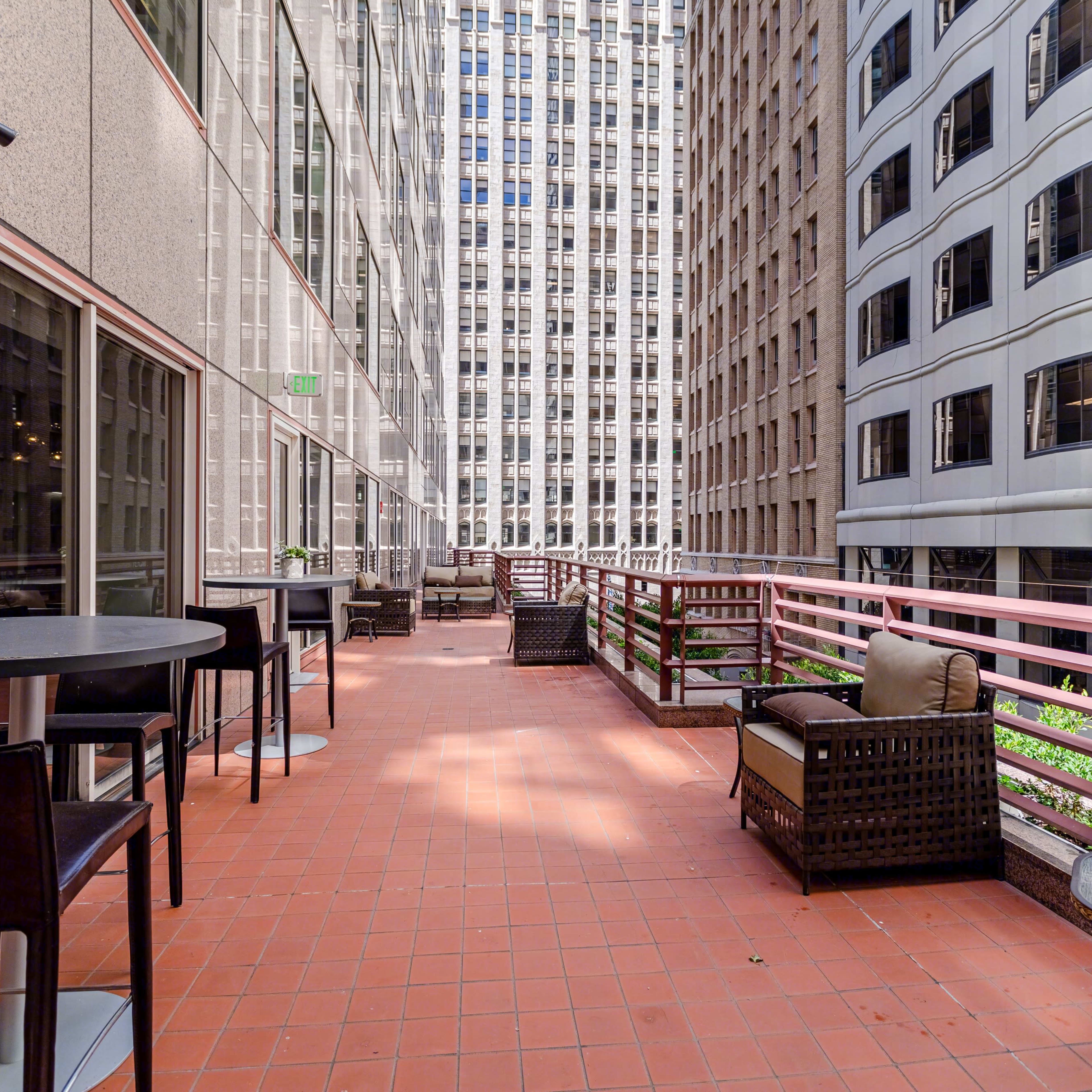 A balcony area with several tables and chairs overlooking a narrow atrium between tall buildings.