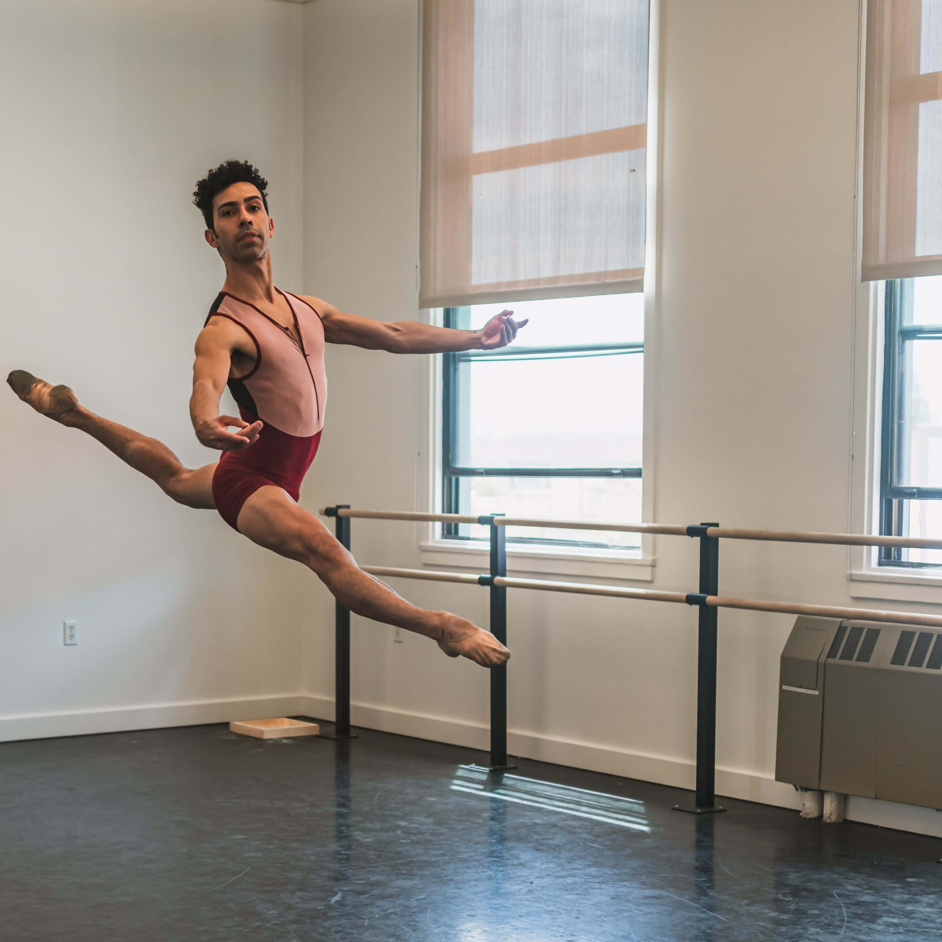 A male ballet dancer performs a leap in a spacious dance studio with large windows and a wooden floor.