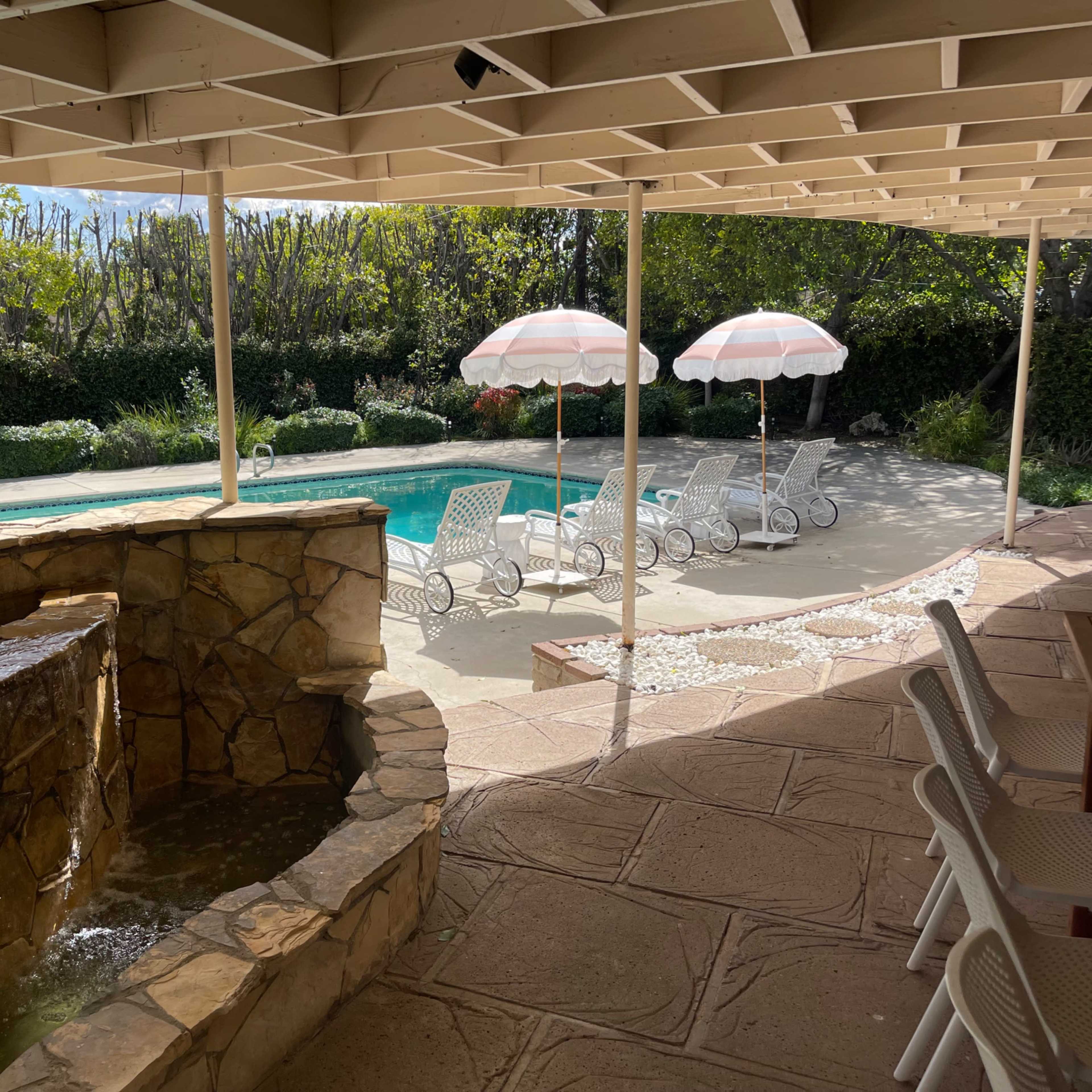 A pool area with a stone fountain in the foreground, white lounge chairs, and two pink umbrellas near the water.