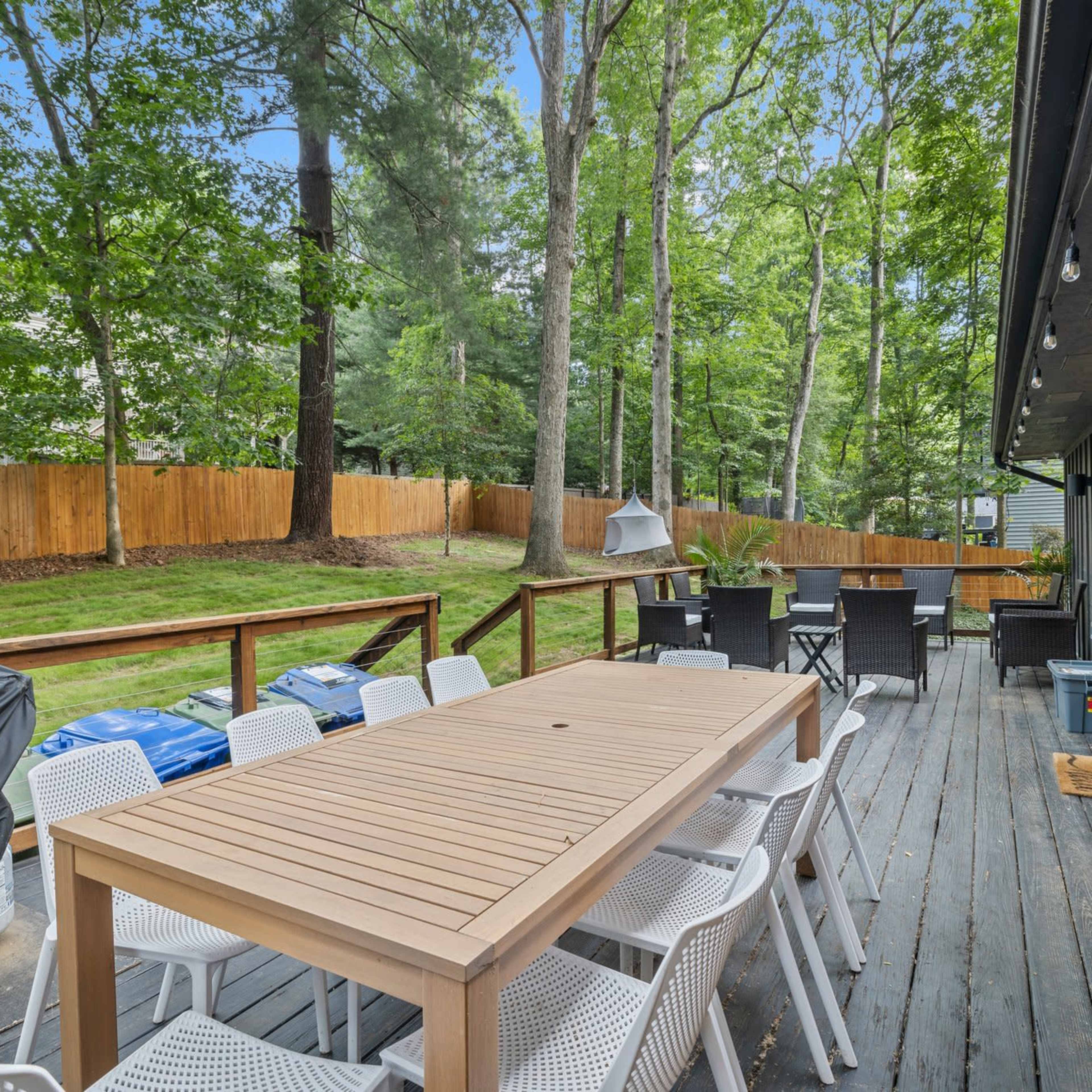 A wooden deck features a long table surrounded by white chairs, with a grill and lush green trees in the background.