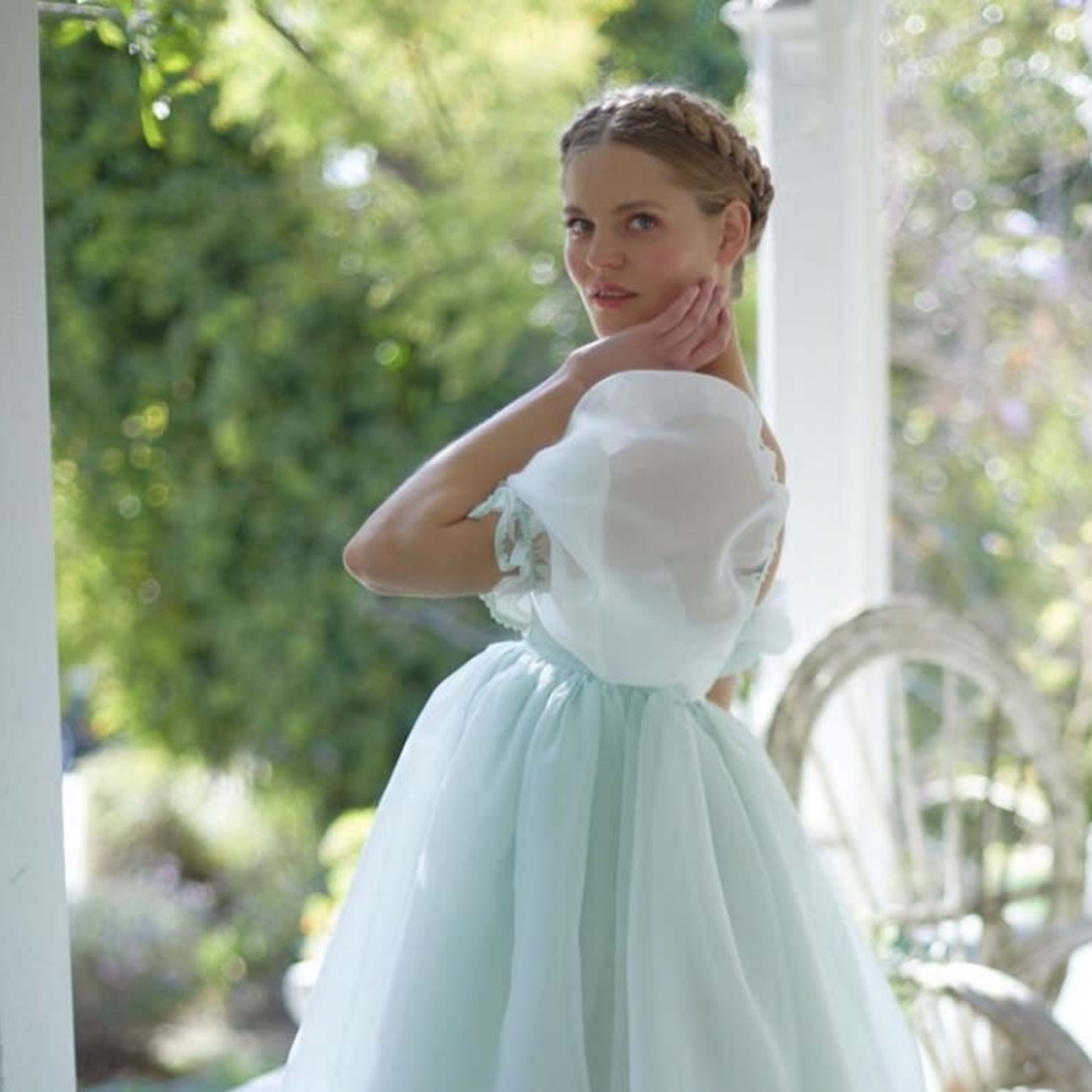 A woman in a light blue dress poses outdoors near a porch surrounded by greenery.