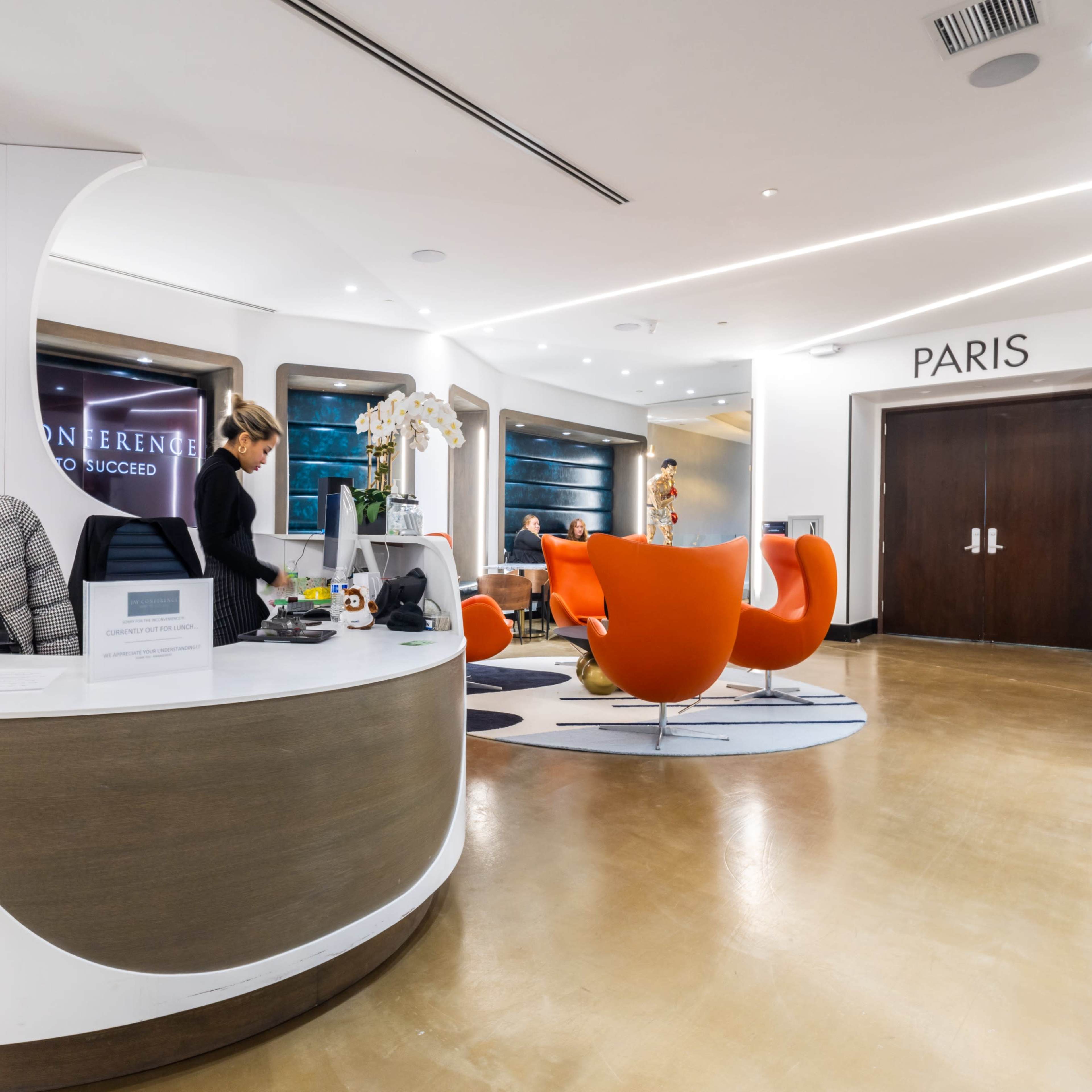 A modern reception area featuring a curved front desk, orange chairs, and a sign indicating "PARIS" on the right.