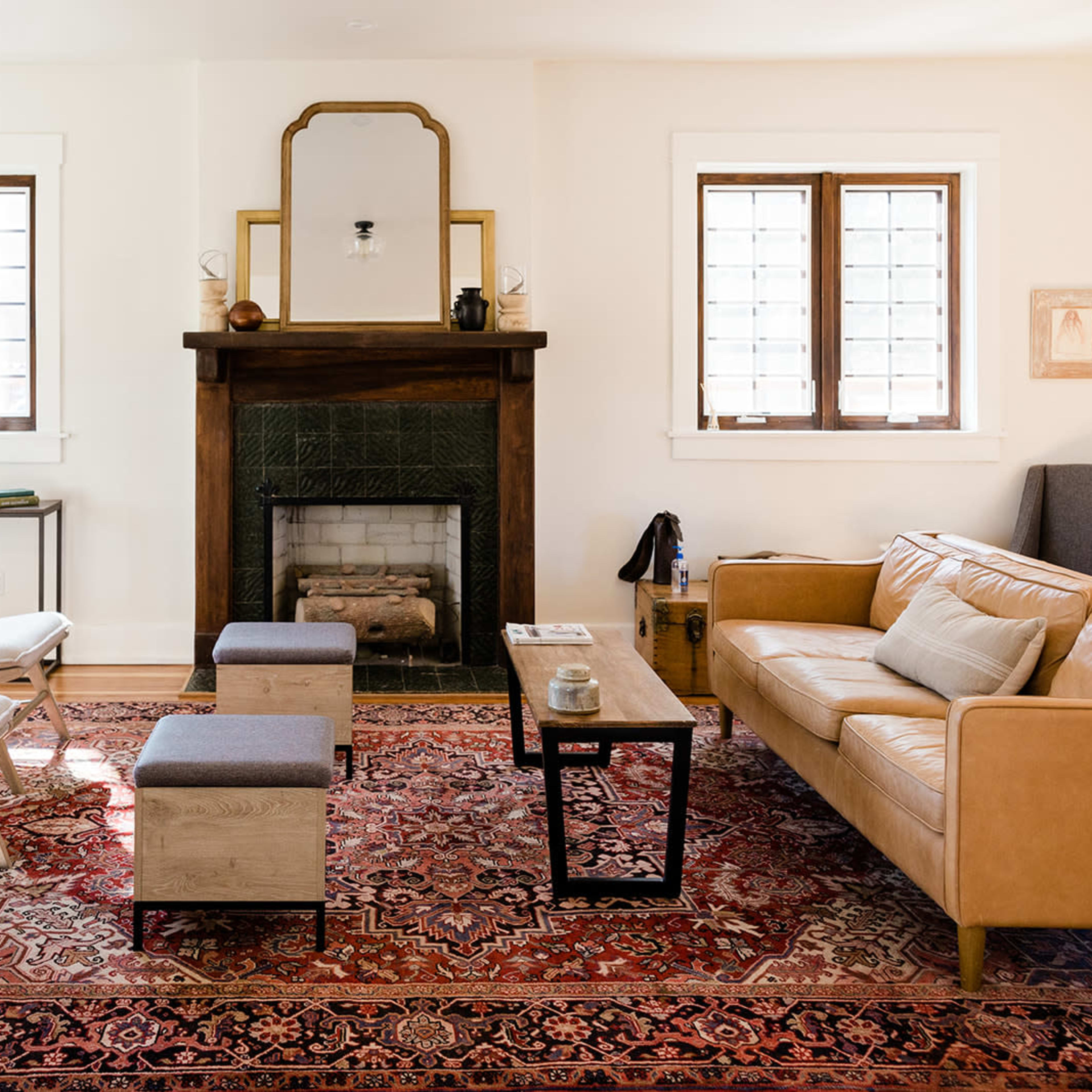 The room features a cozy living area with a leather sofa, two armchairs, and a coffee table arranged around a patterned rug, with a fireplace and windows providing natural light.