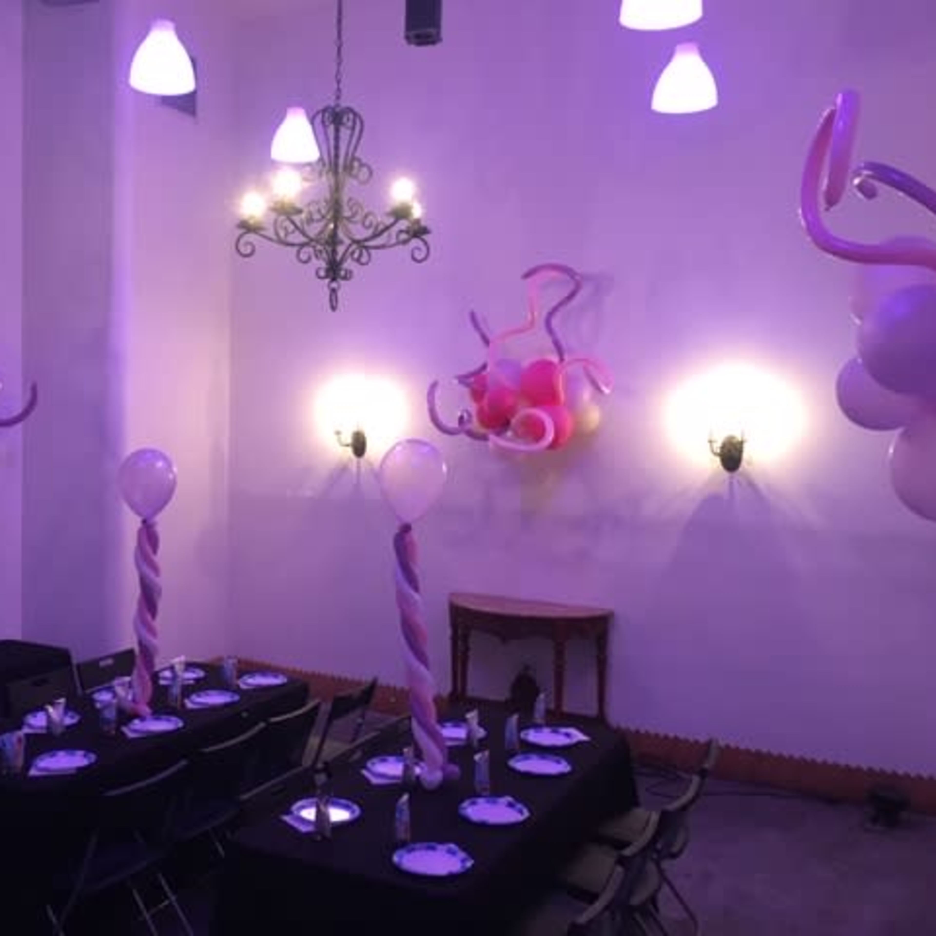 A decorated party room features a black table set for guests, complete with balloons and swirl decorations in shades of pink.