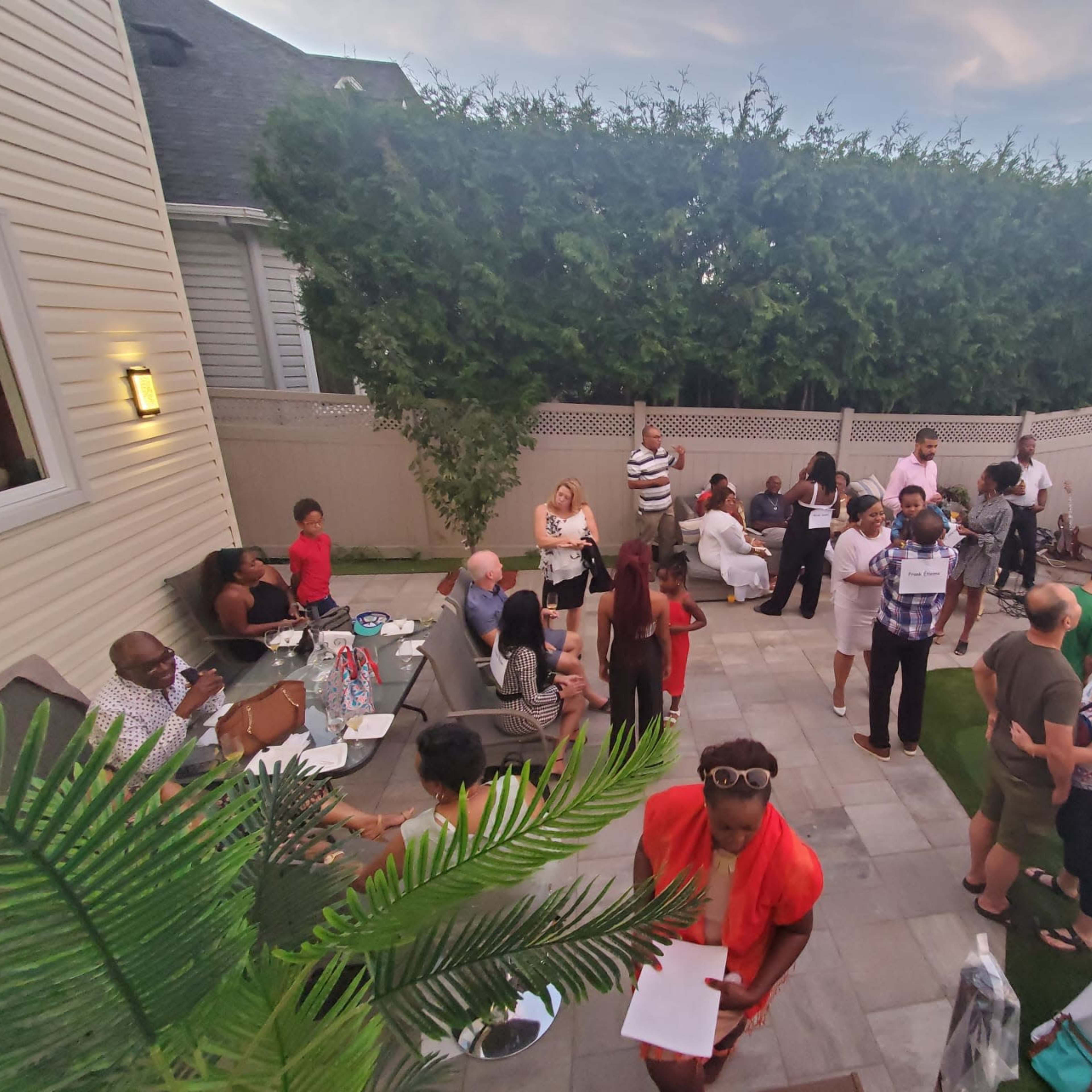 A group of people is gathered in a backyard, engaging in conversation and activities around seating areas and tables.