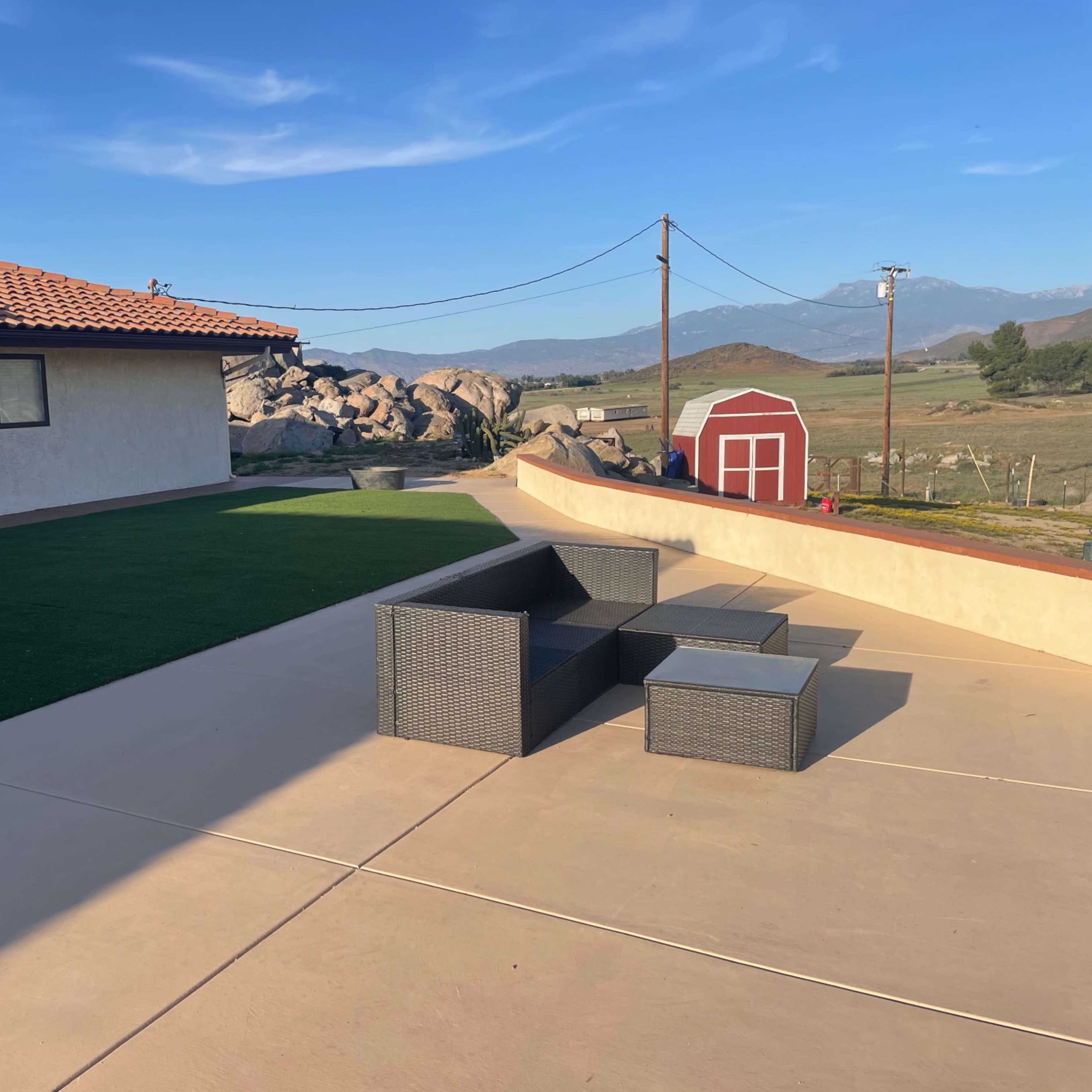 The scene features a patio area with a dark wicker seating set, a grassy section, and a red barn in the background against a mountain range.