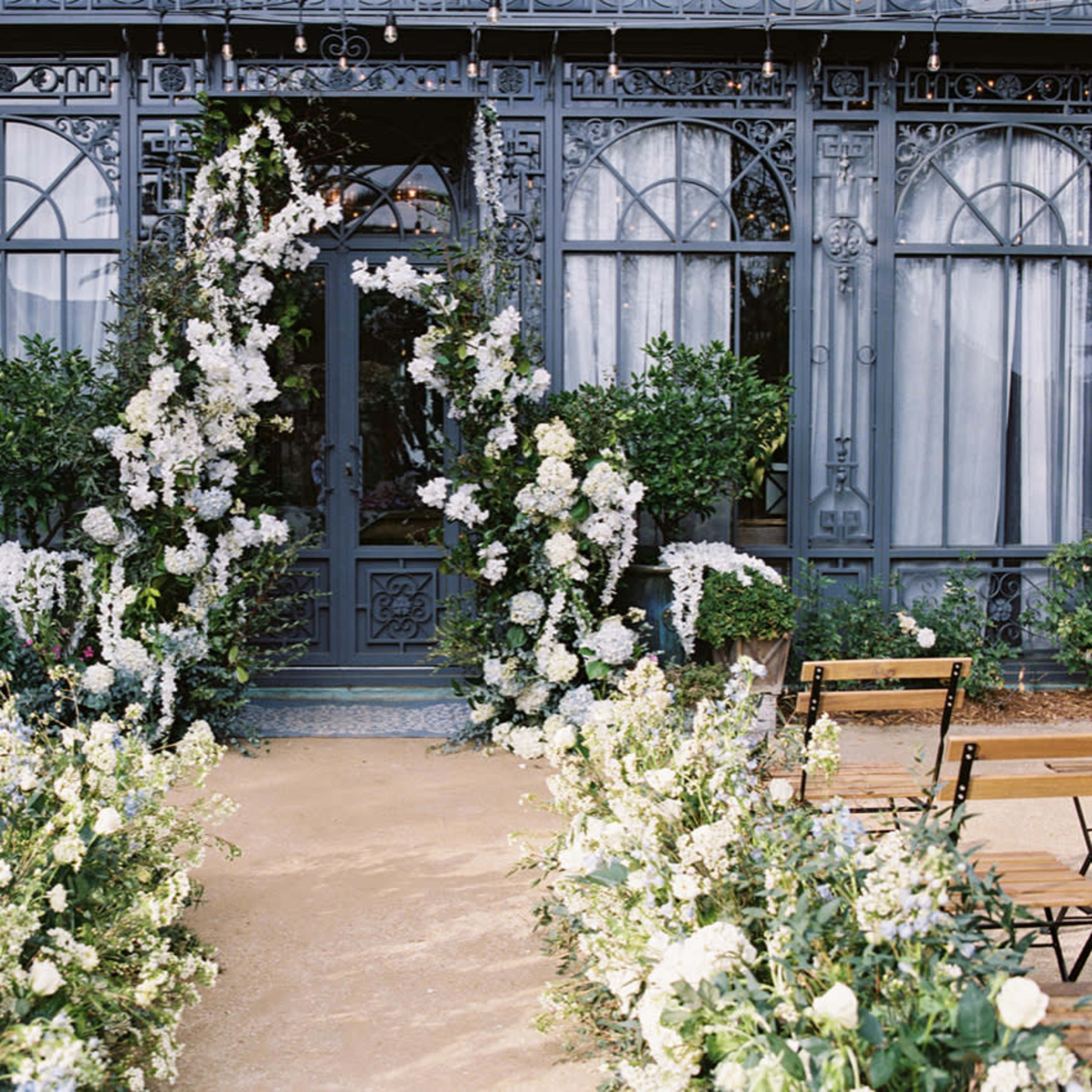 An elegantly decorated entrance featuring a doorway framed by lush white flowers and green foliage, with wooden chairs arranged in lines leading towards it.