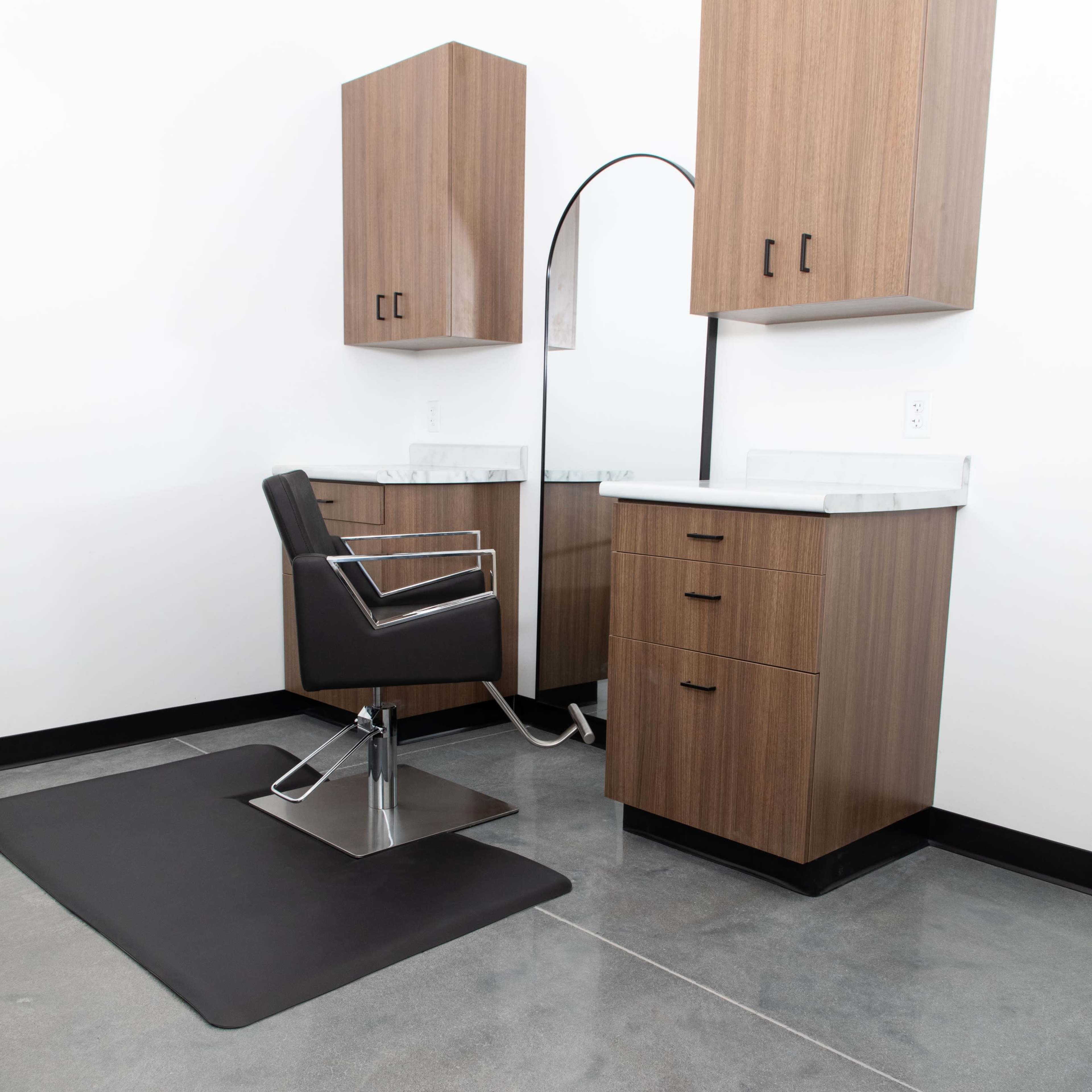 A modern hair salon setup features a black styling chair, a large mirror, and wooden cabinetry against a white wall.