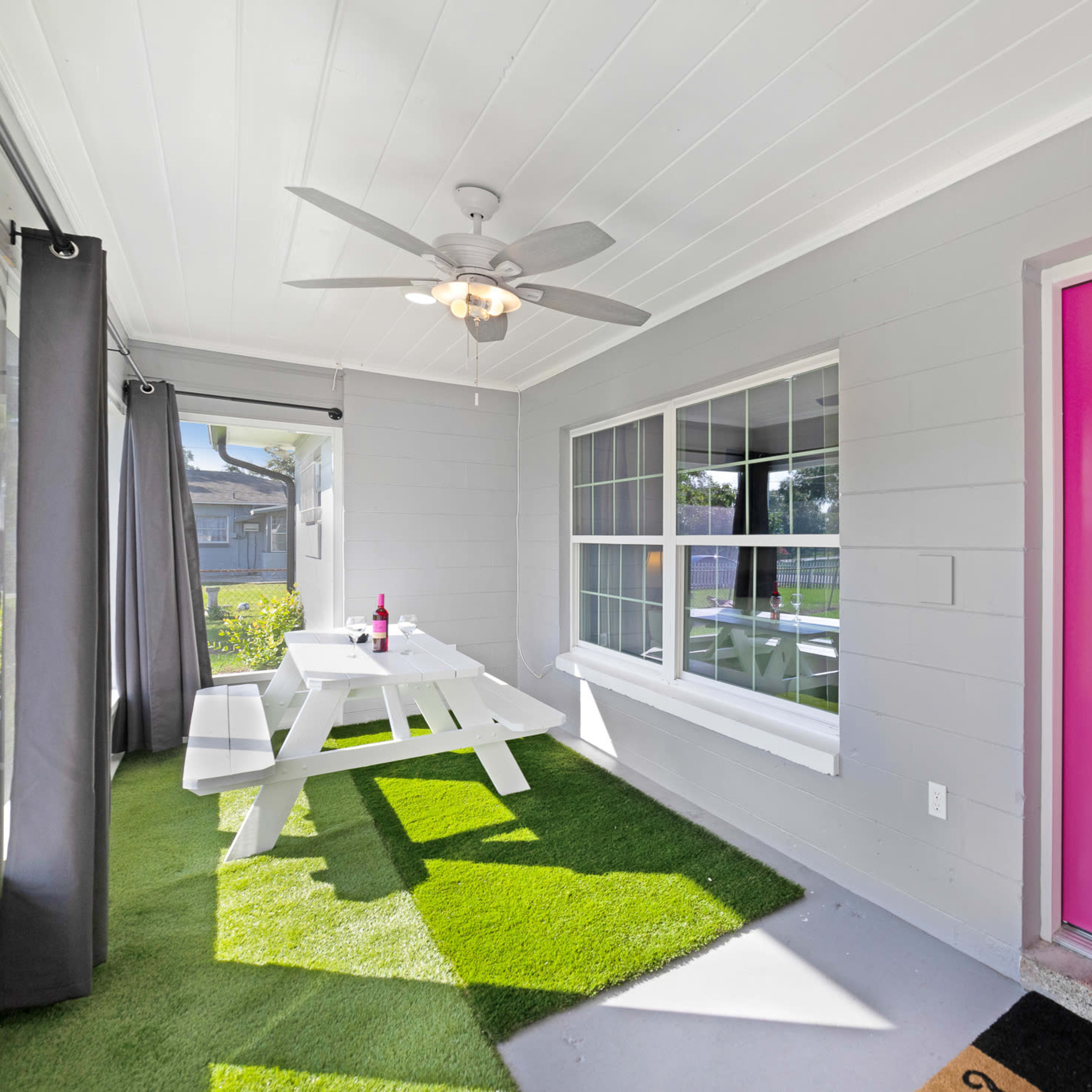 A bright pink door stands out against a light gray exterior, leading to a porch with a picnic table and artificial grass.