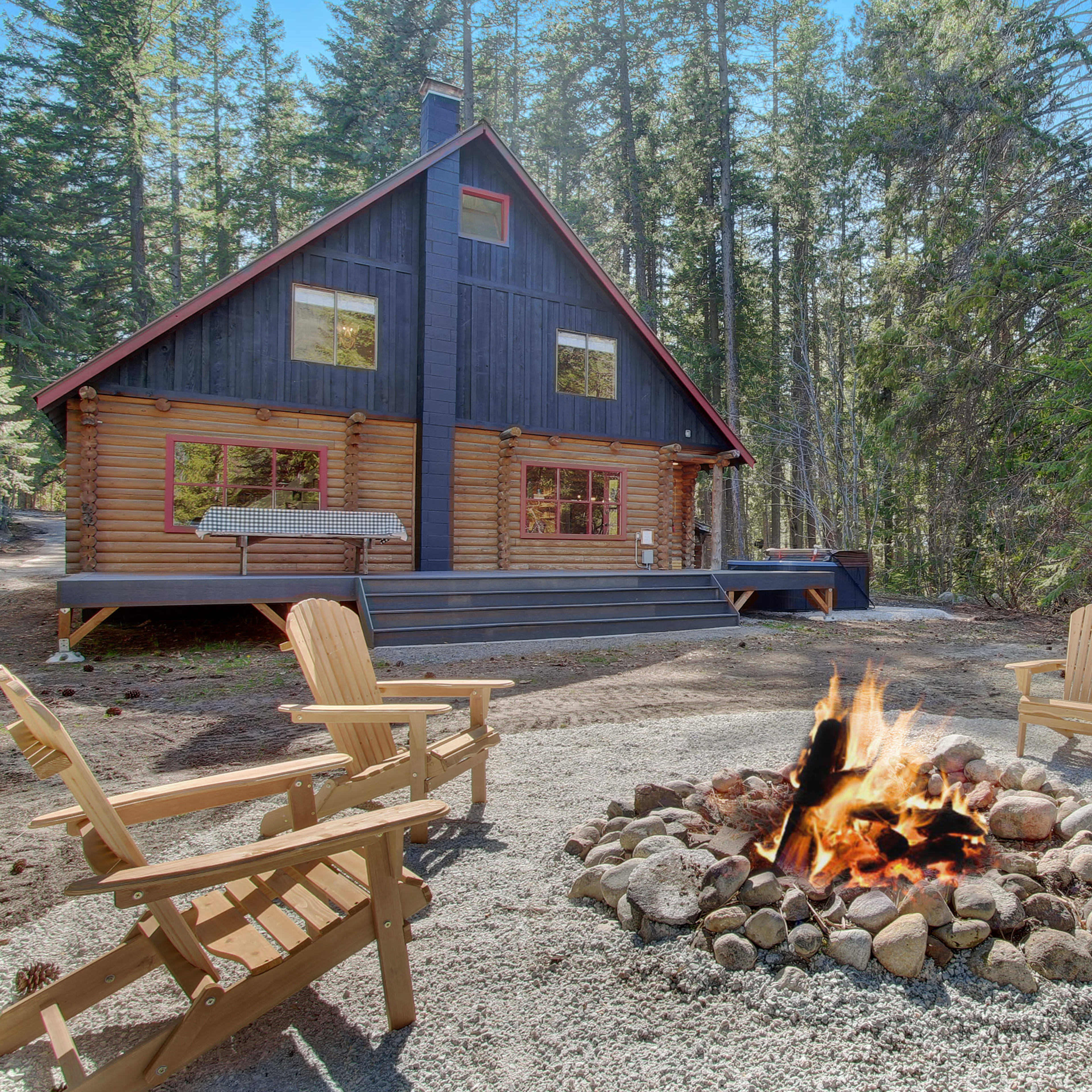 A log cabin with a red roof is surrounded by trees, featuring a circular stone fire pit with four wooden chairs in front.