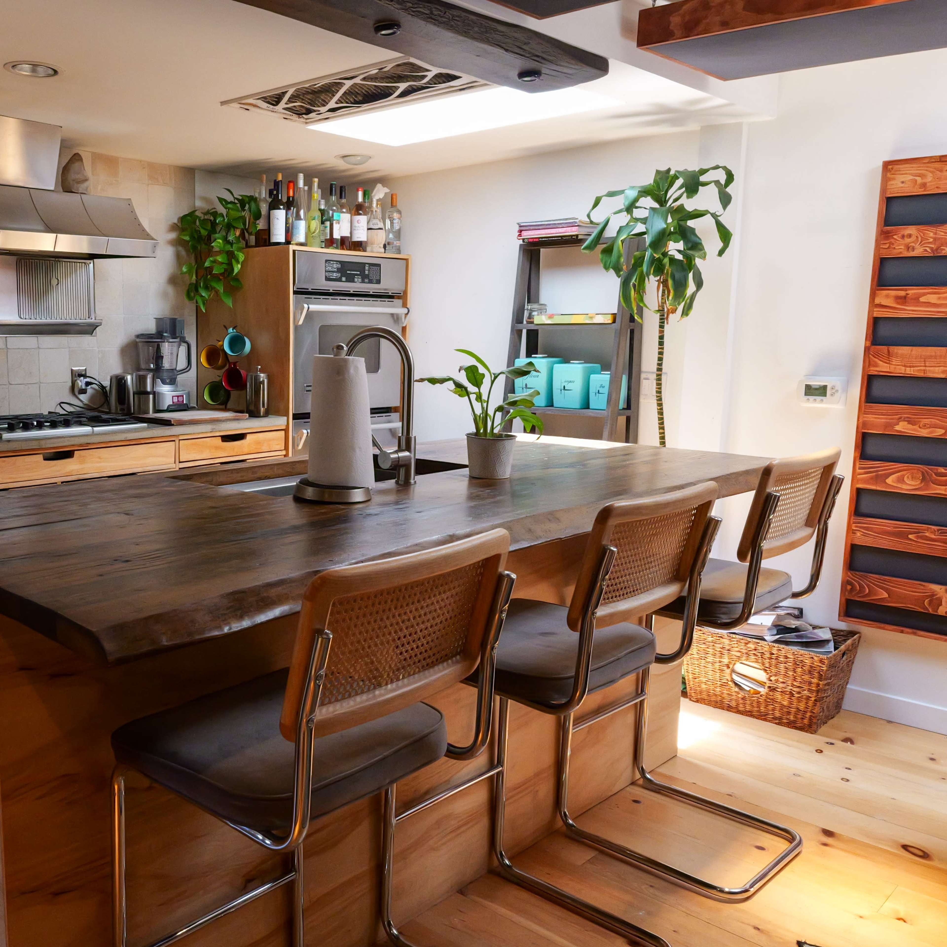 A modern kitchen features a wooden island with bar stools, stainless steel appliances, and potted plants in a well-lit space.
