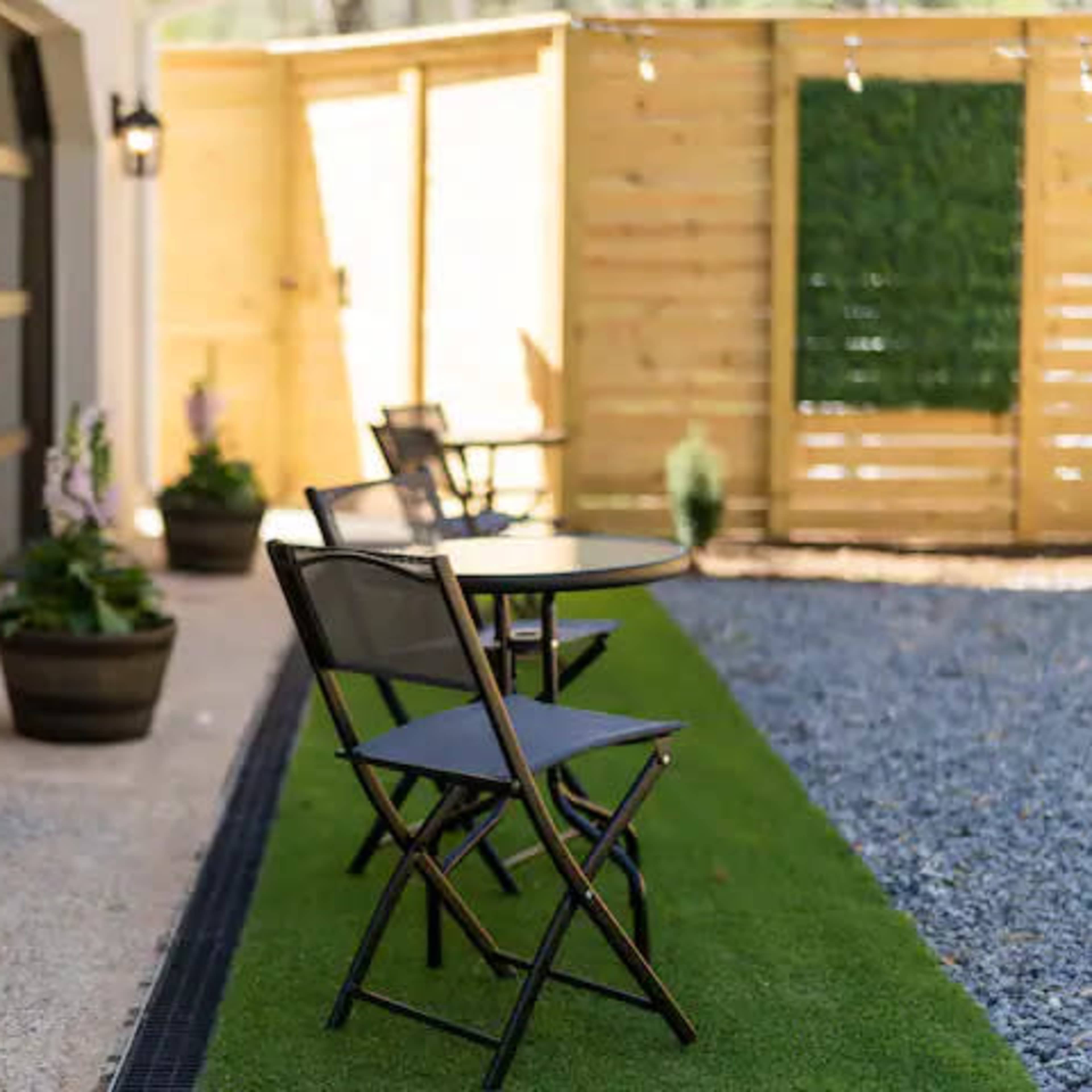 A small outdoor seating area features a round table and two folding chairs on artificial grass, bordered by potted plants and a wooden privacy fence.