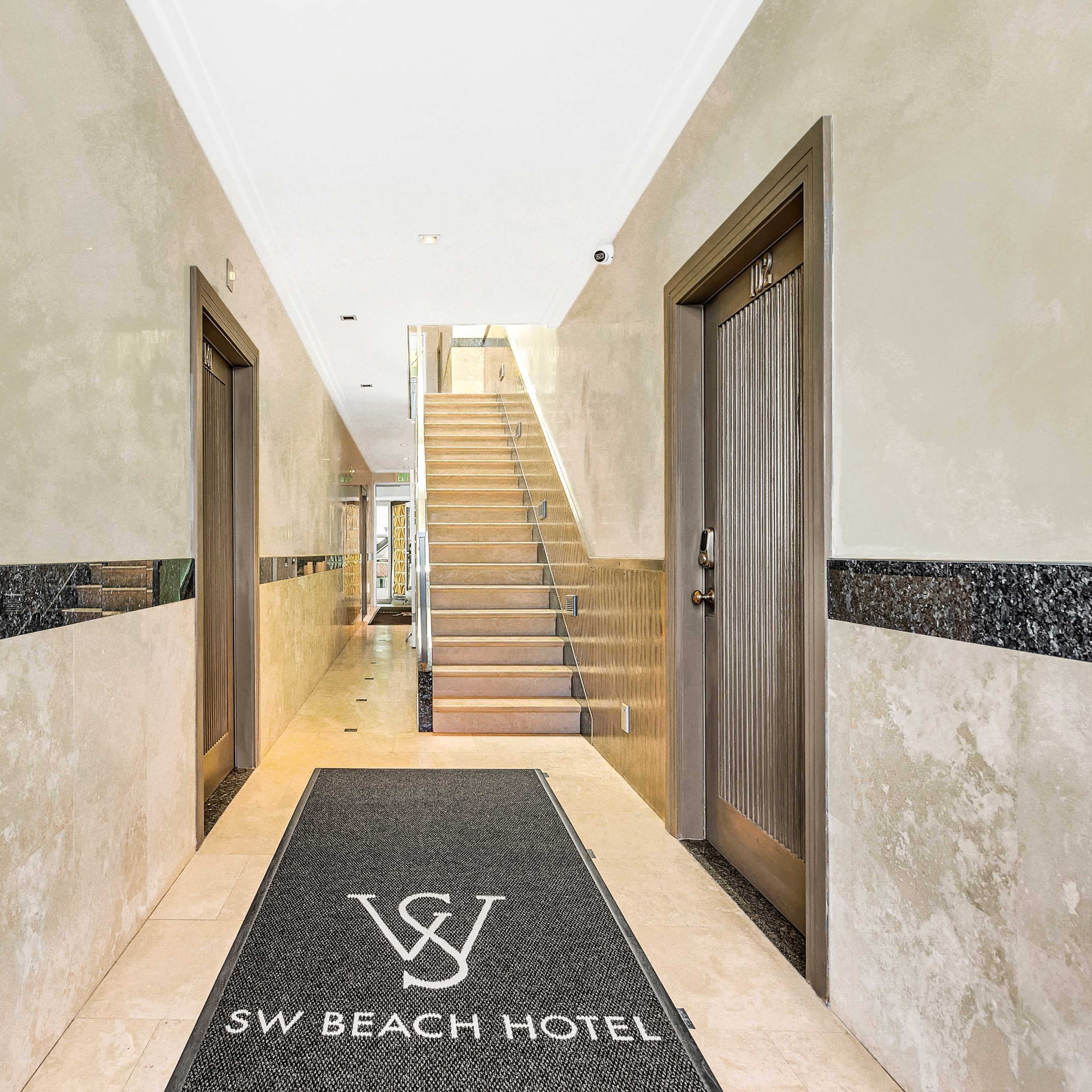 A hallway leading to a set of stairs with doors on either side, featuring a logo for "SW Beach Hotel" on a floor mat.