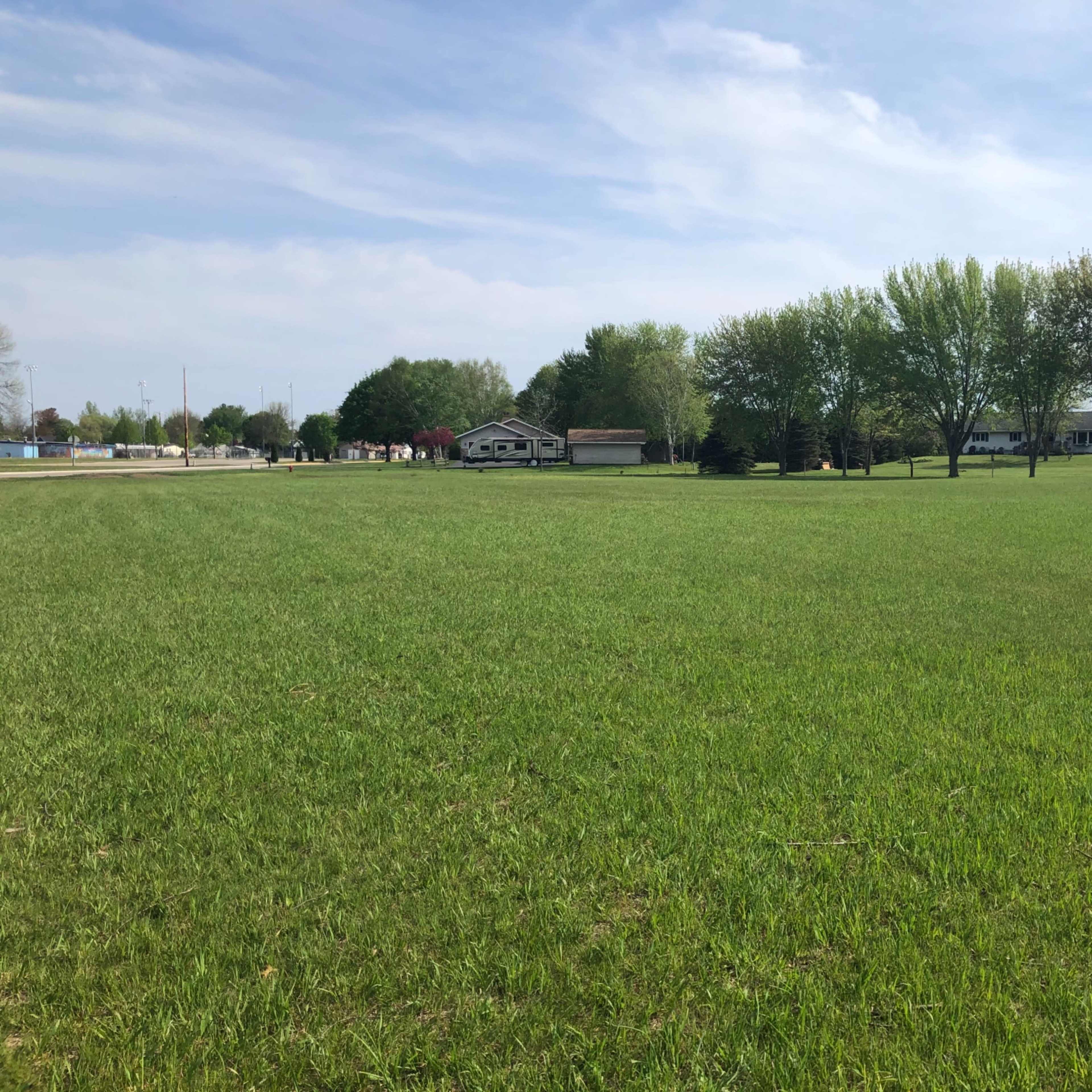 A vast green field with a few trees and houses lining the background under a partly cloudy sky.