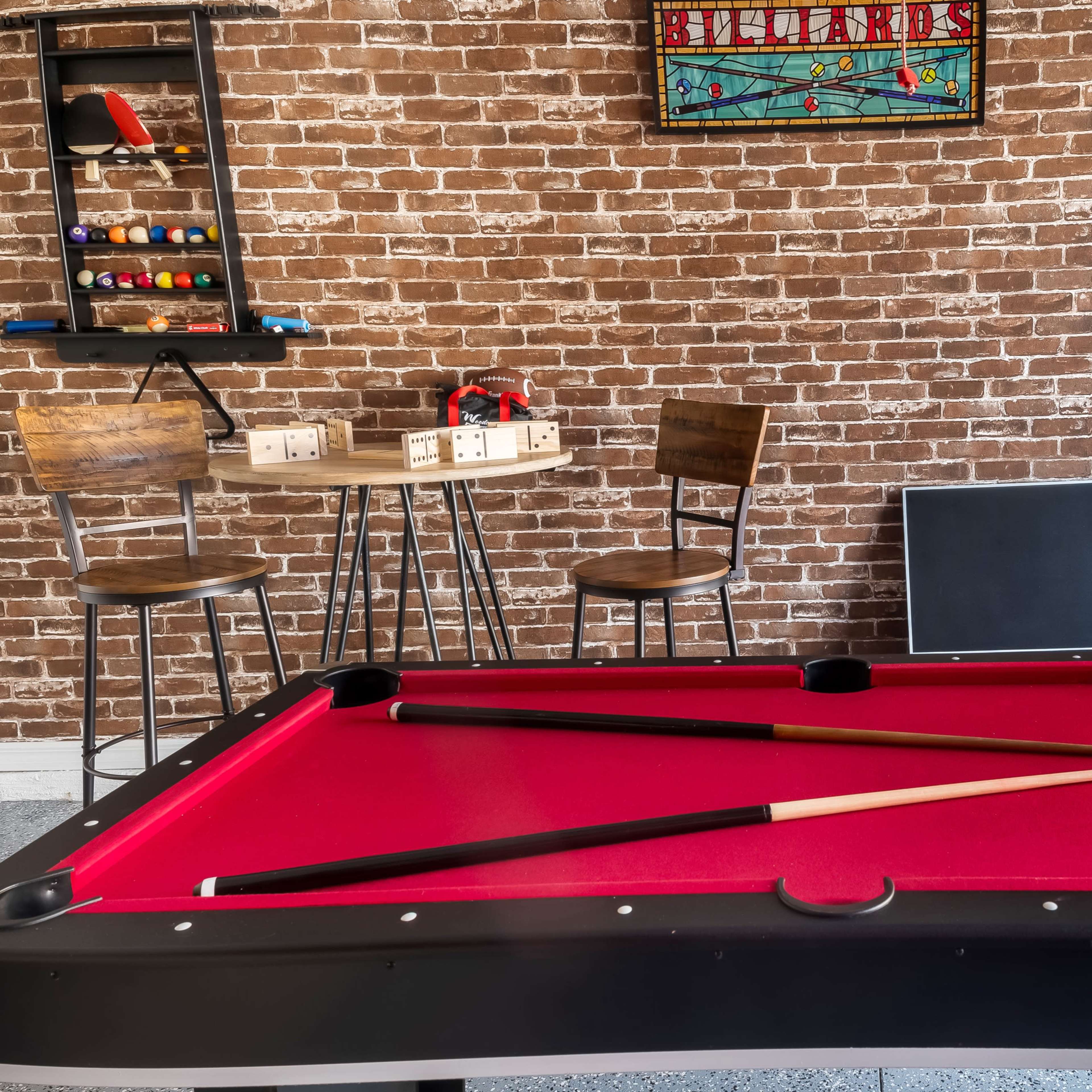 A red-felt billiard table is centered in a room with a brick wall, featuring a small table and chairs in the background.