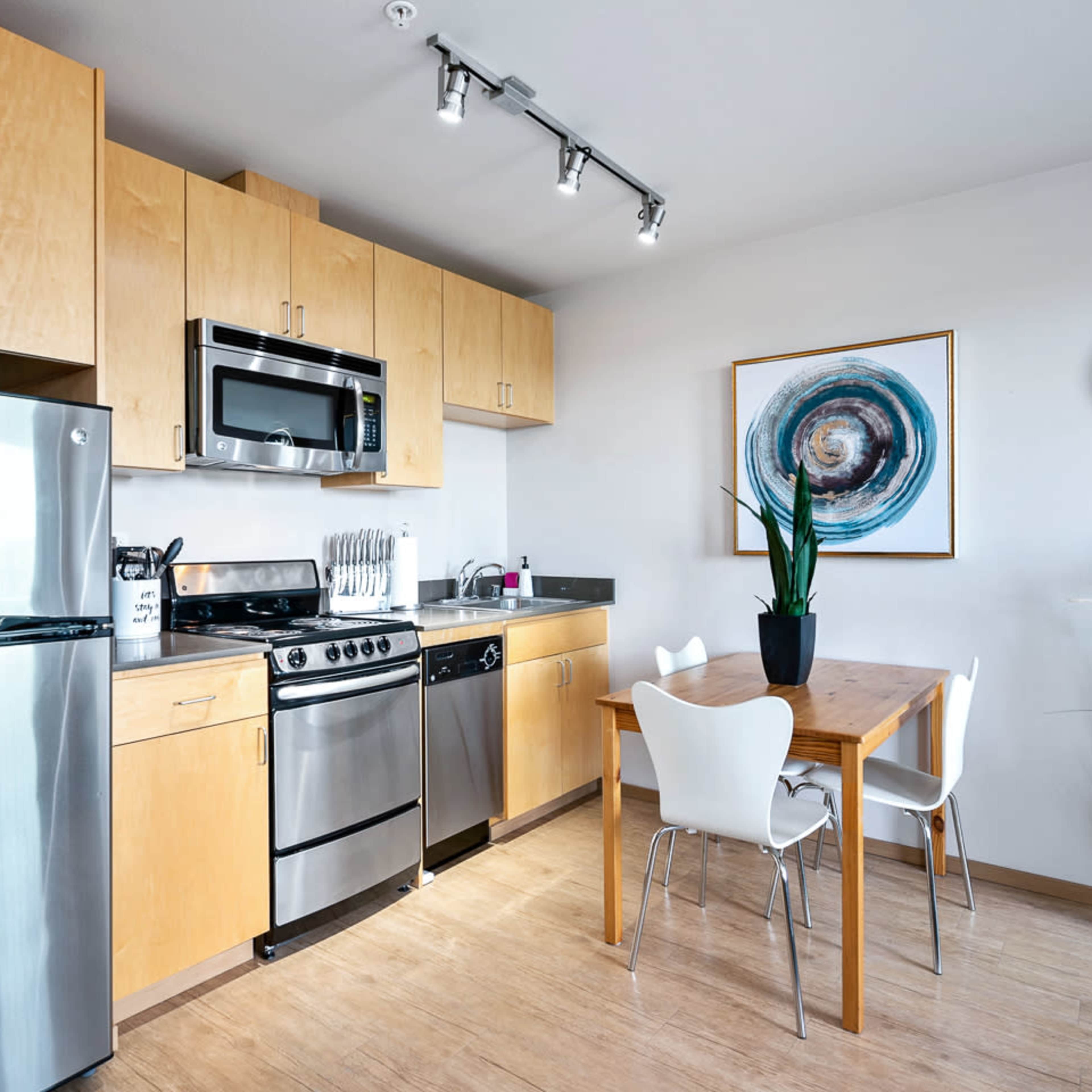 A modern kitchen area featuring stainless steel appliances, wooden cabinets, a dining table with white chairs, and a decorative plant.