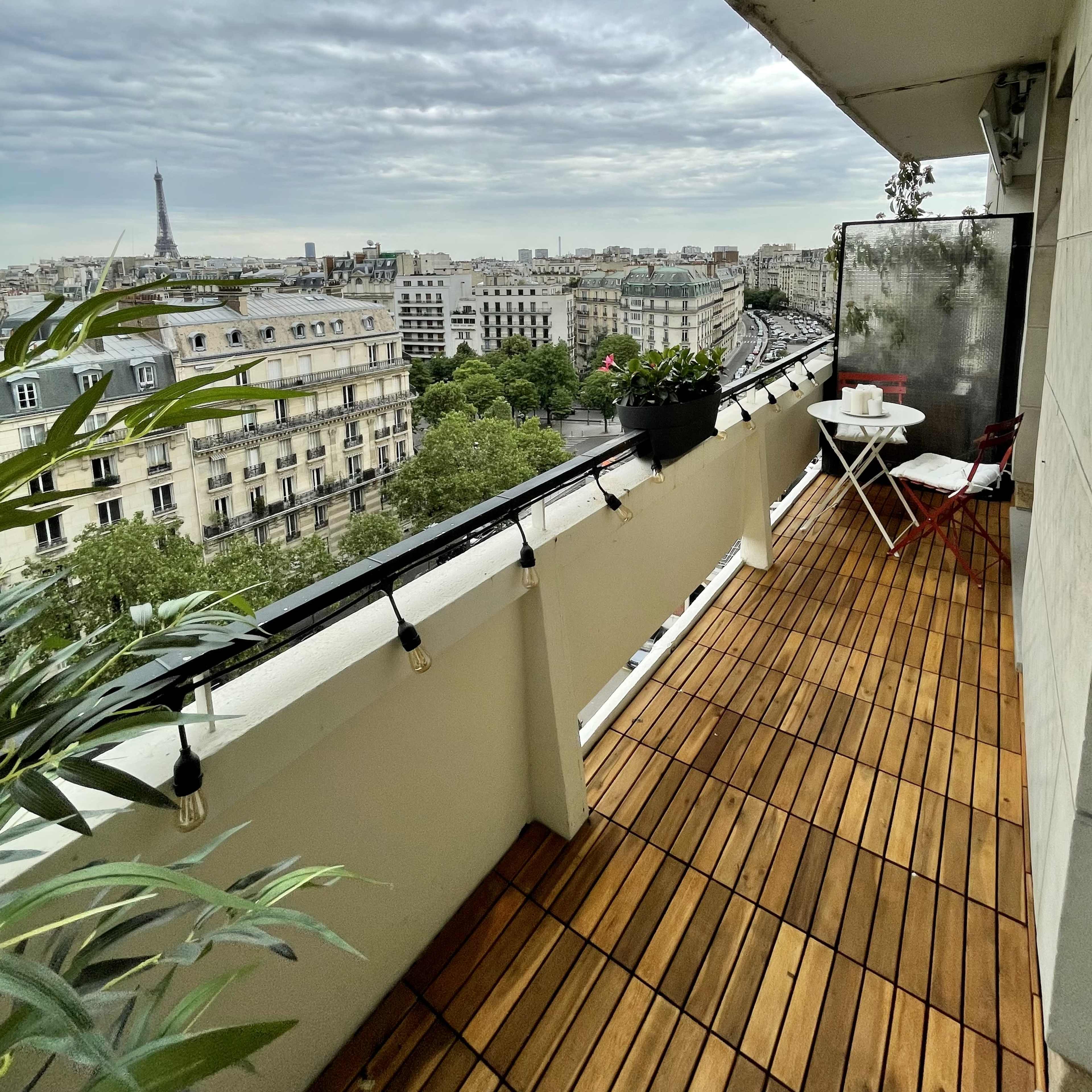 A balcony with wooden flooring features a small table and chairs, overlooking a cityscape that includes the Eiffel Tower.
