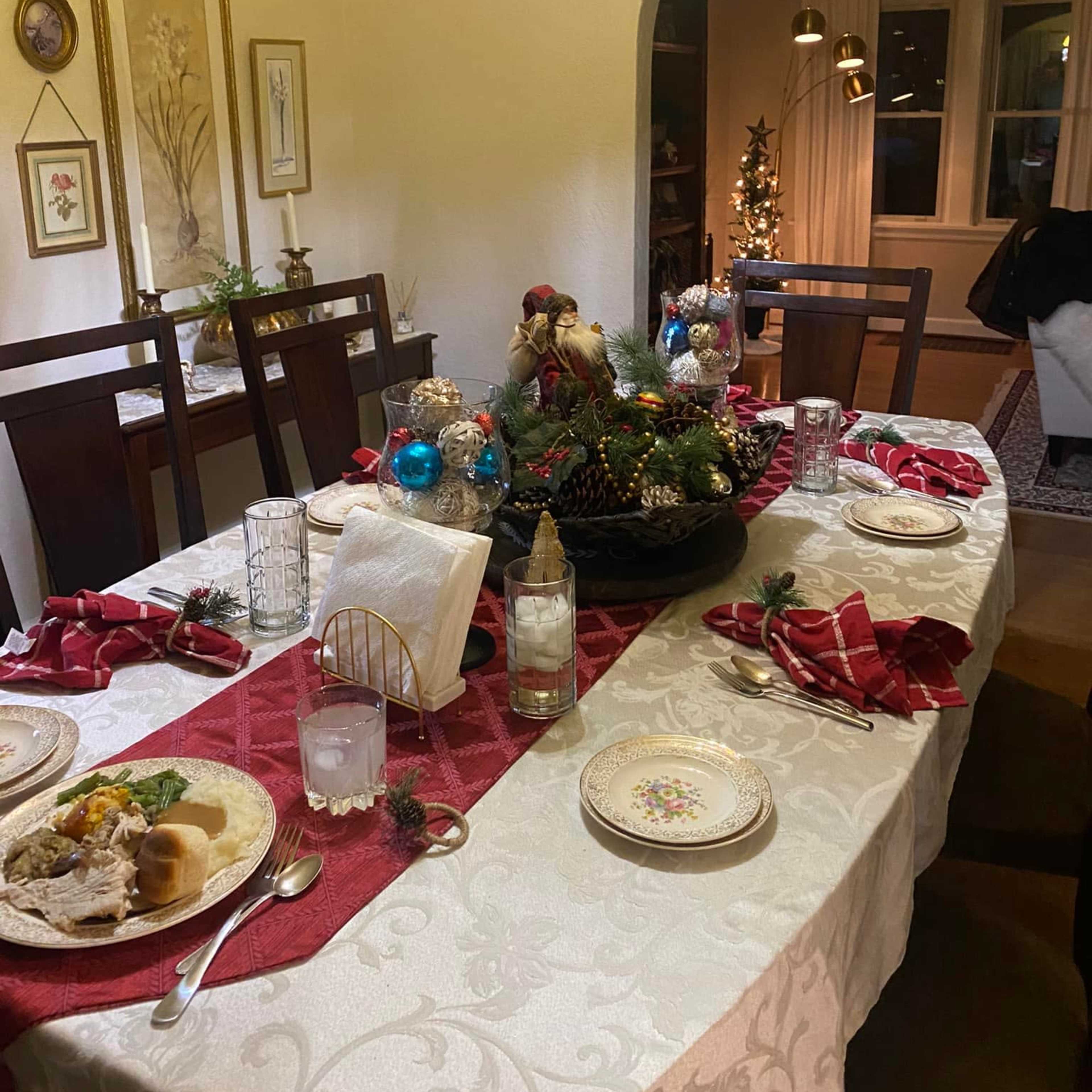 A dining table is set for a holiday meal, featuring decorative elements, plates, and glasses, with a Christmas tree visible in the background.