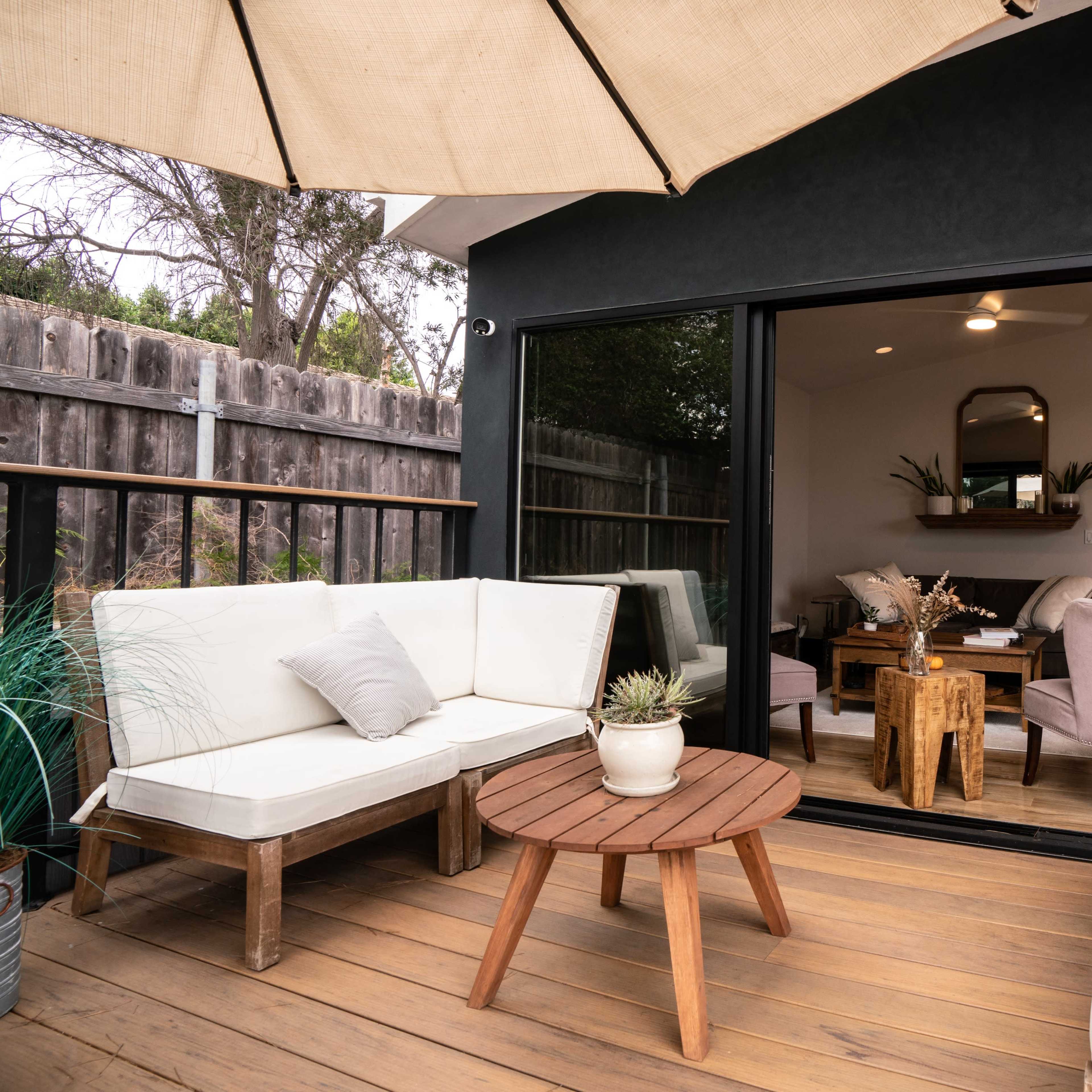 A modern patio features a seating area with a wooden coffee table, an umbrella, and a view into a well-lit interior space.