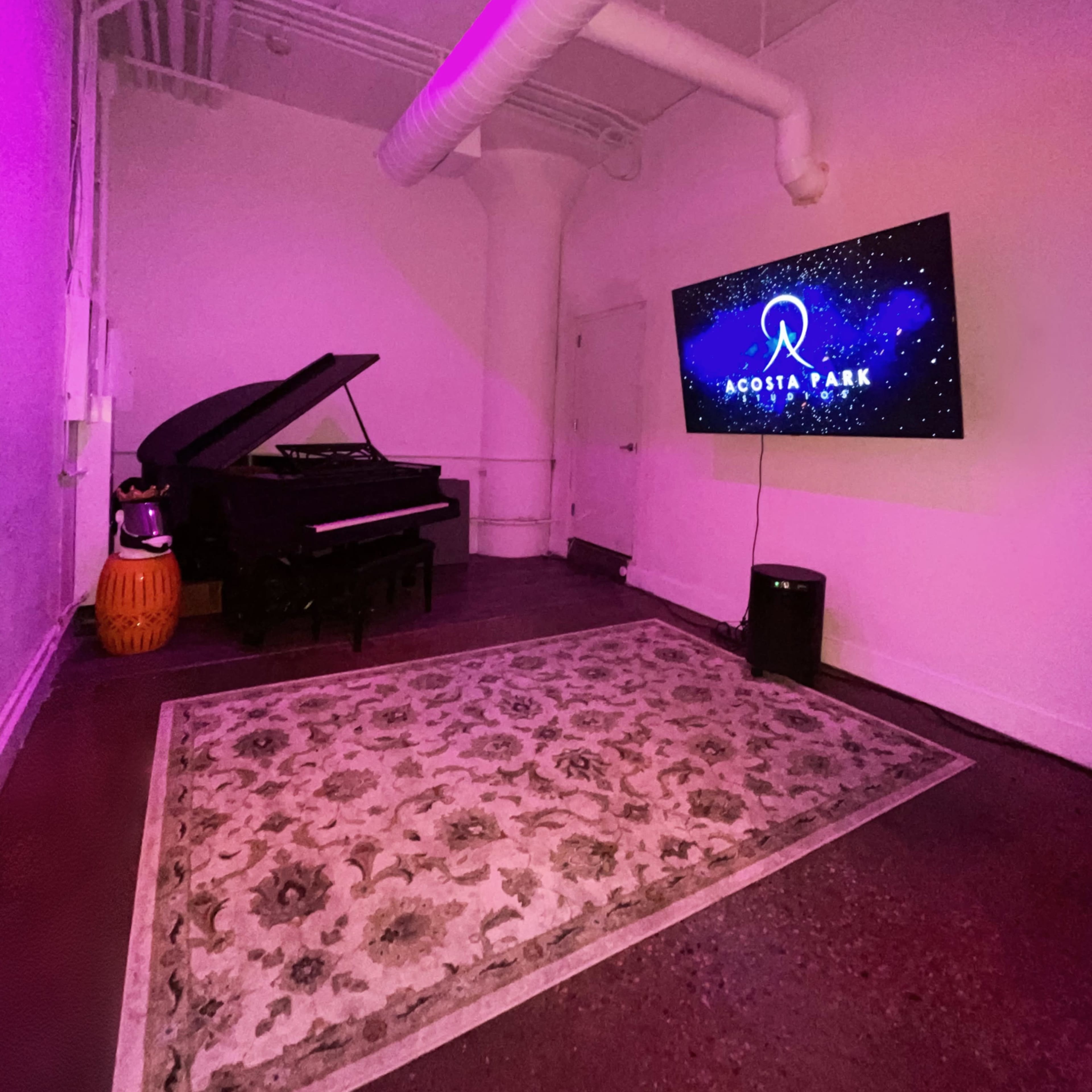 A grand piano is positioned next to a television displaying the logo for "ACOSTA PARK" in a room with purple lighting and a decorative rug on the floor.