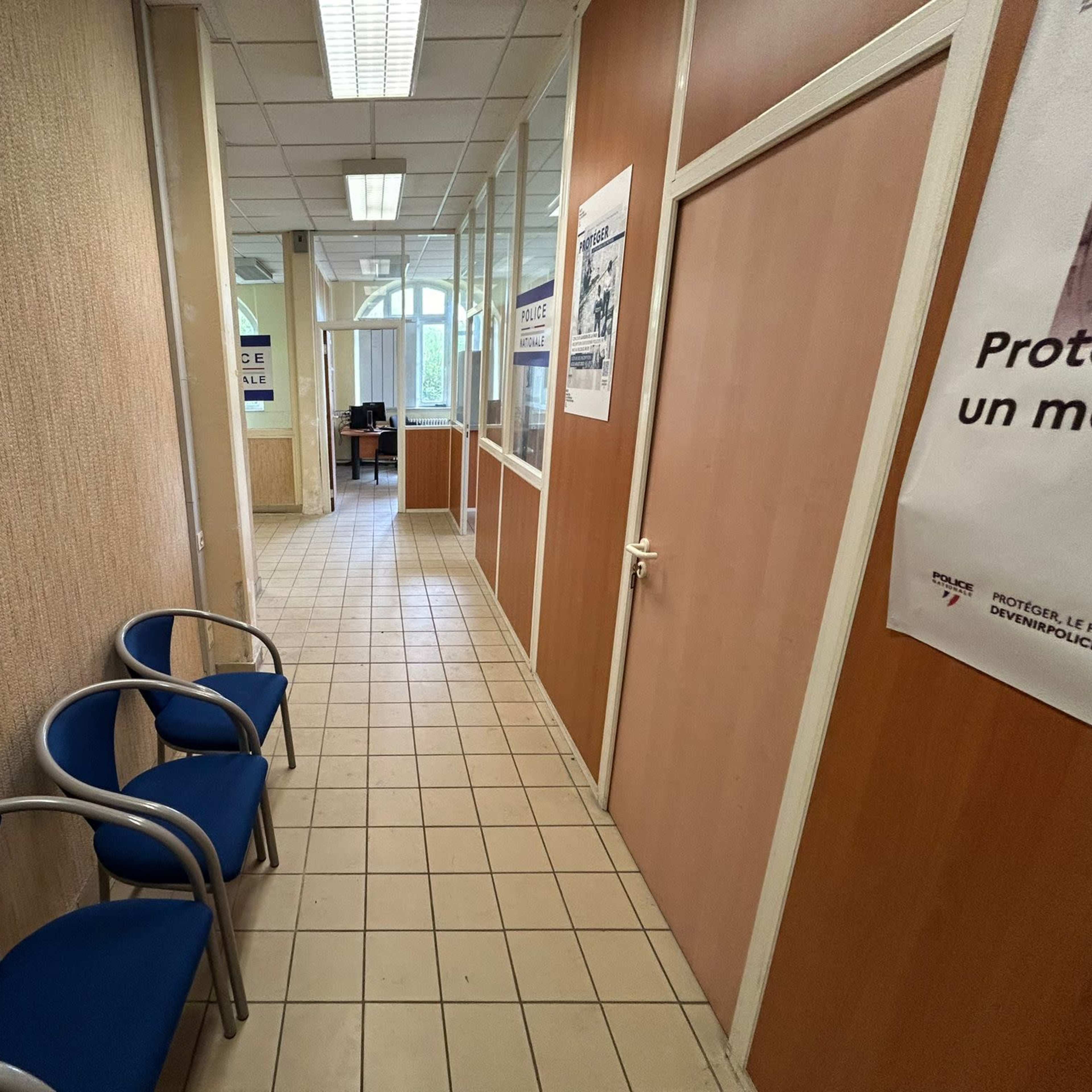 A narrow corridor with tile flooring, leading to an office at the end, accompanied by blue chairs on the left side and glass-paneled walls.