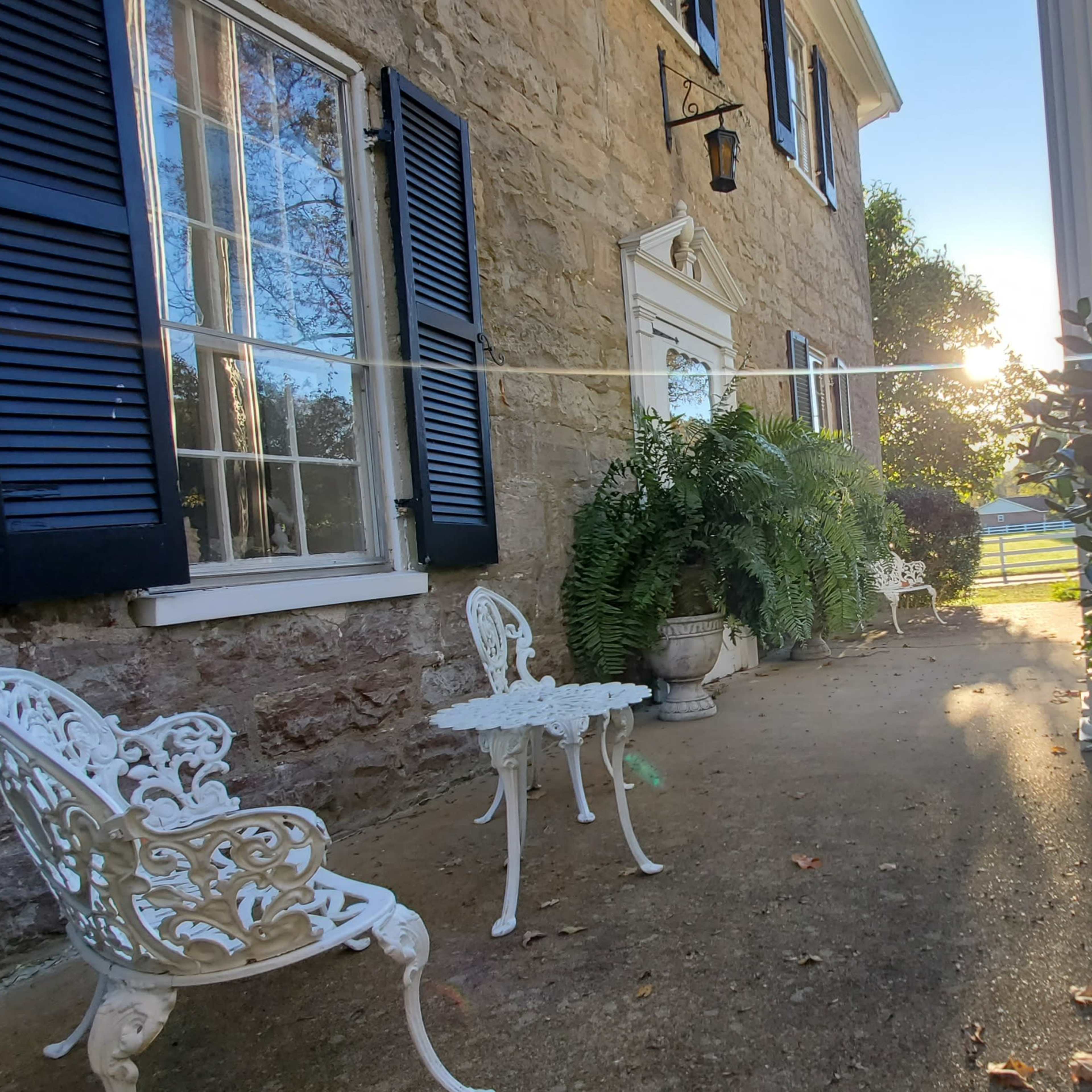 A stone wall encases a pathway lined with chairs and potted plants, with sunlight shining through the trees at the end.