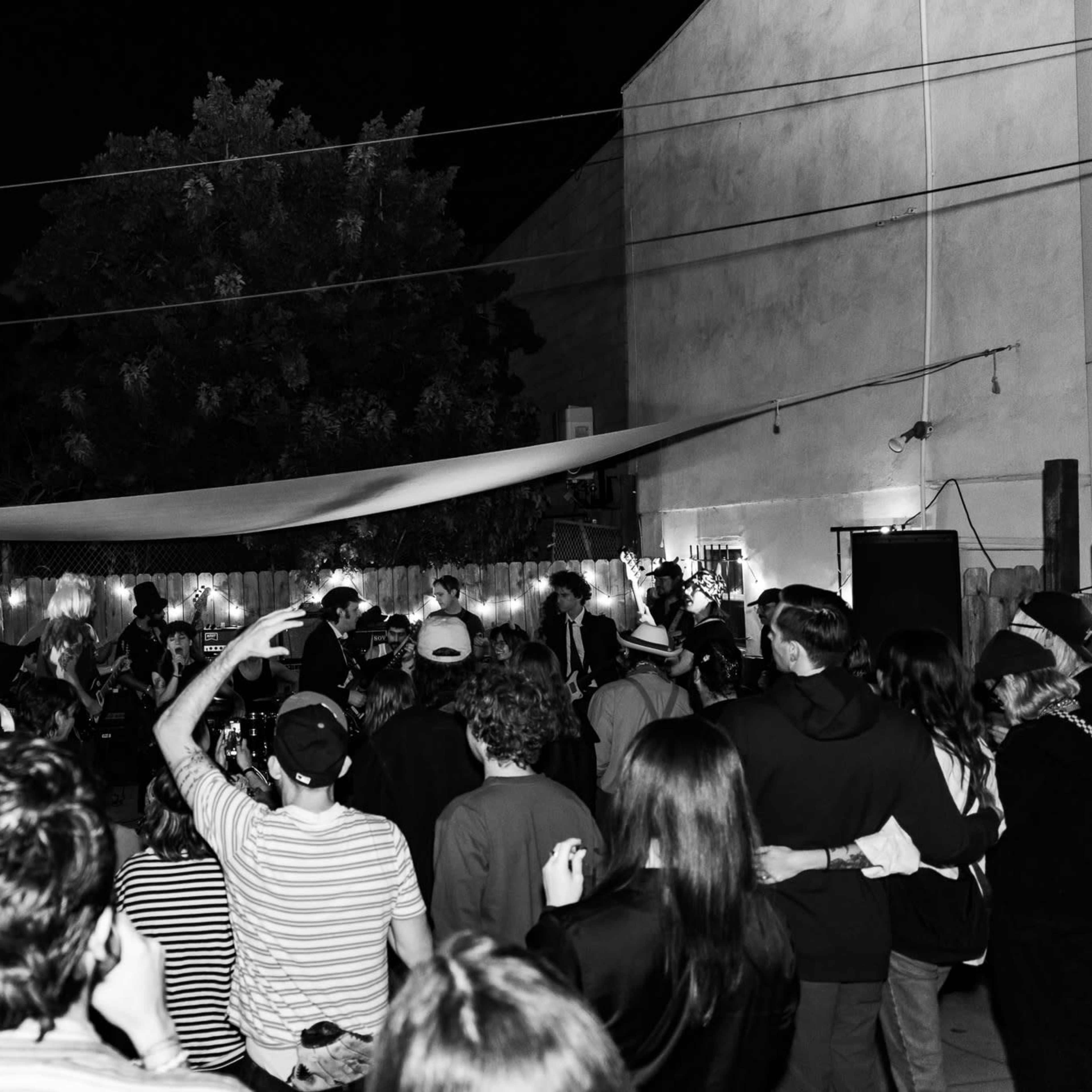 A crowd of people gathers in a dimly lit outdoor space to watch a live band perform.