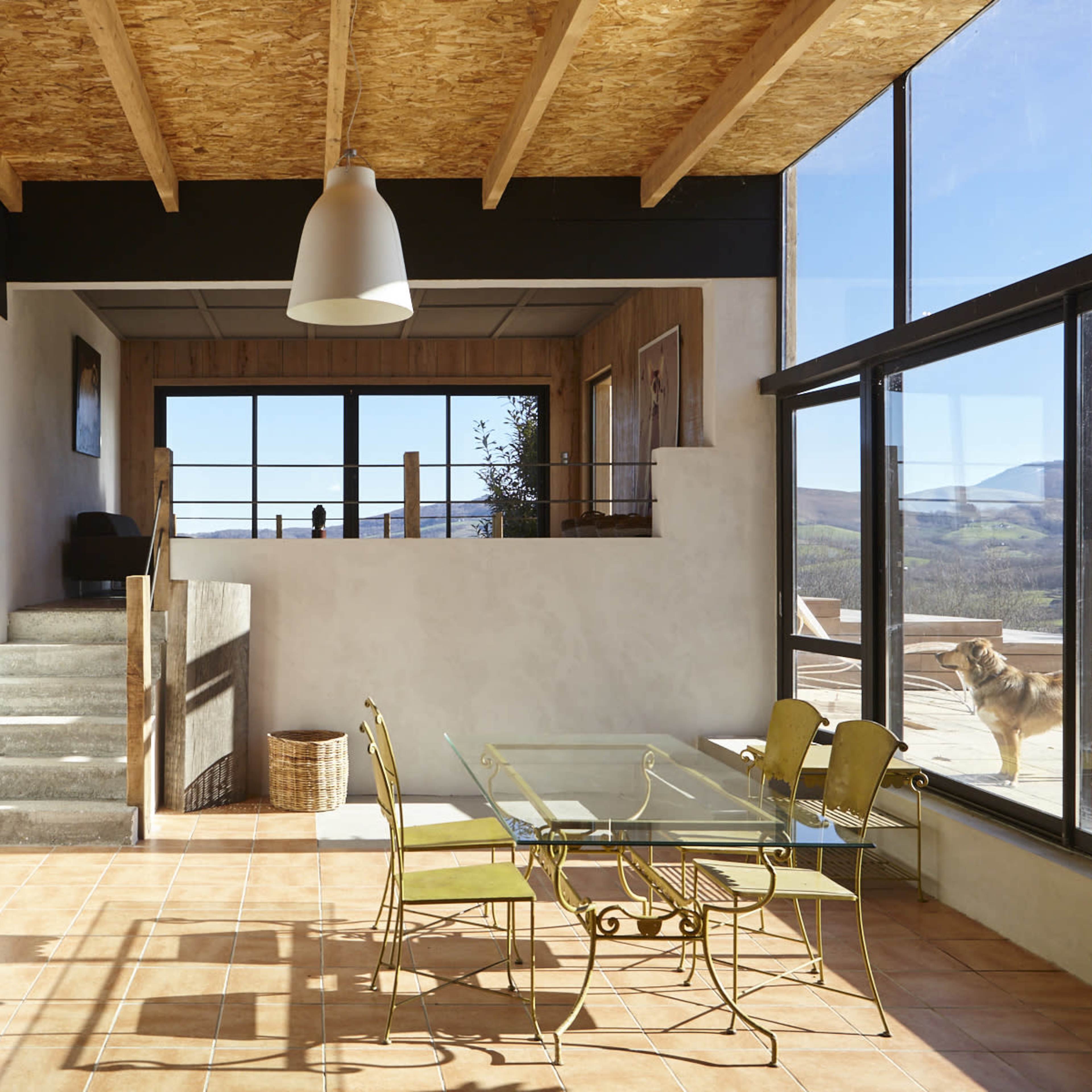 A spacious room with a dining area featuring a glass table and yellow chairs, large windows that allow natural light, and a staircase leading to an upper level.