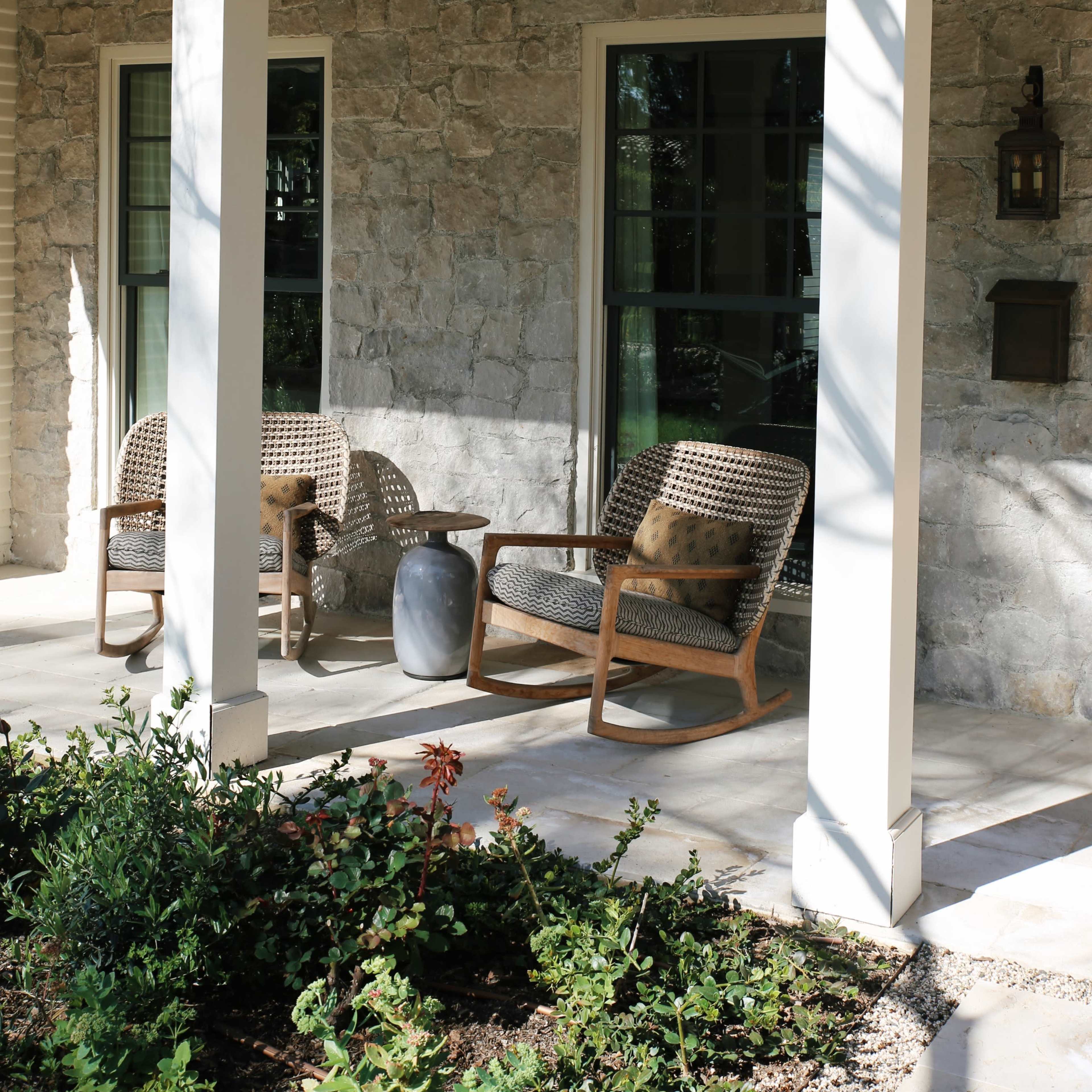 A stone cottage porch with two woven chairs, a small side table, and a lantern mounted on the wall.