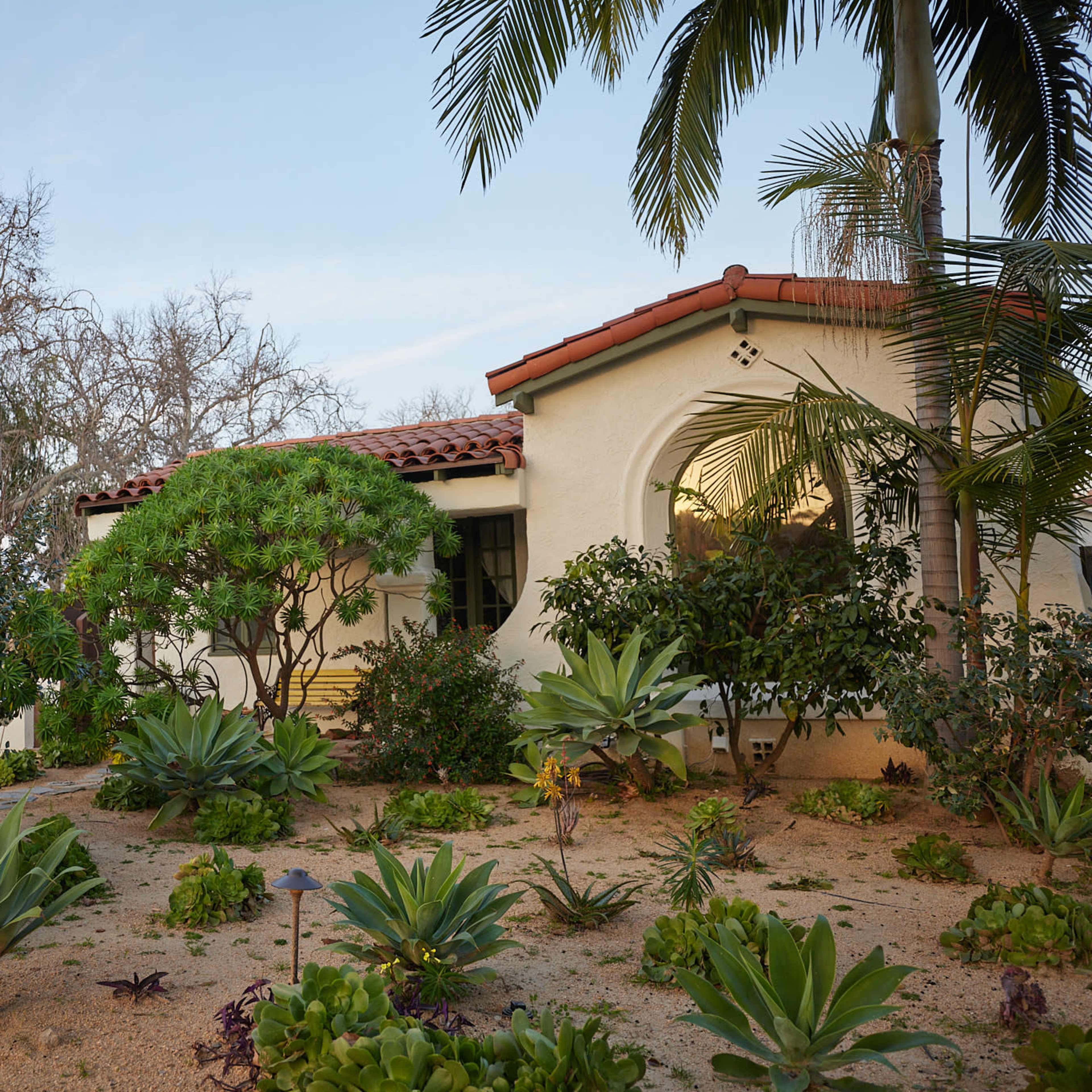 A white stucco house with a red-tiled roof is surrounded by a garden featuring various plants and shrubs.