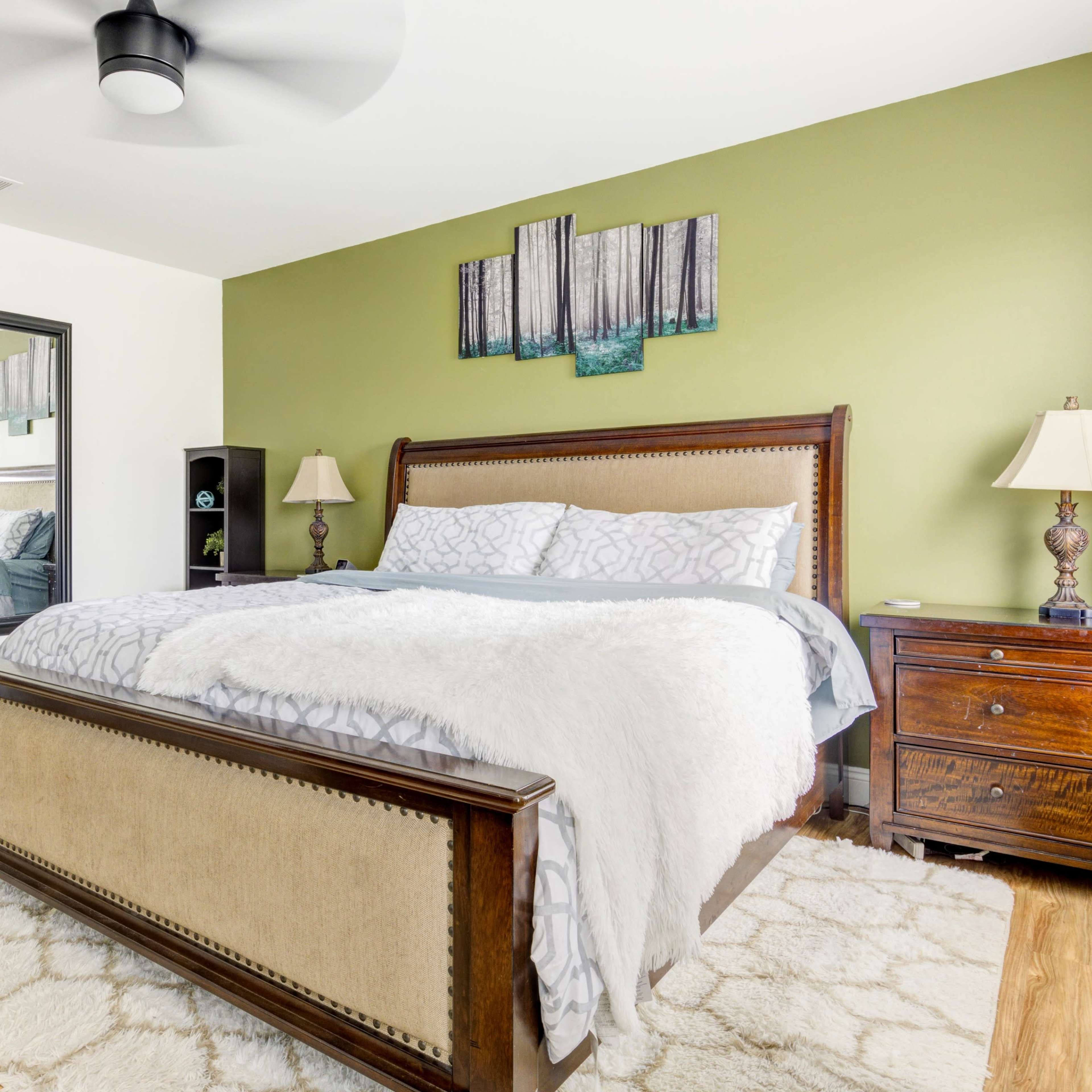 A bedroom features a large bed with a patterned comforter, a wooden nightstand, and green walls adorned with artwork, along with a mirror and an open door leading to a bathroom.