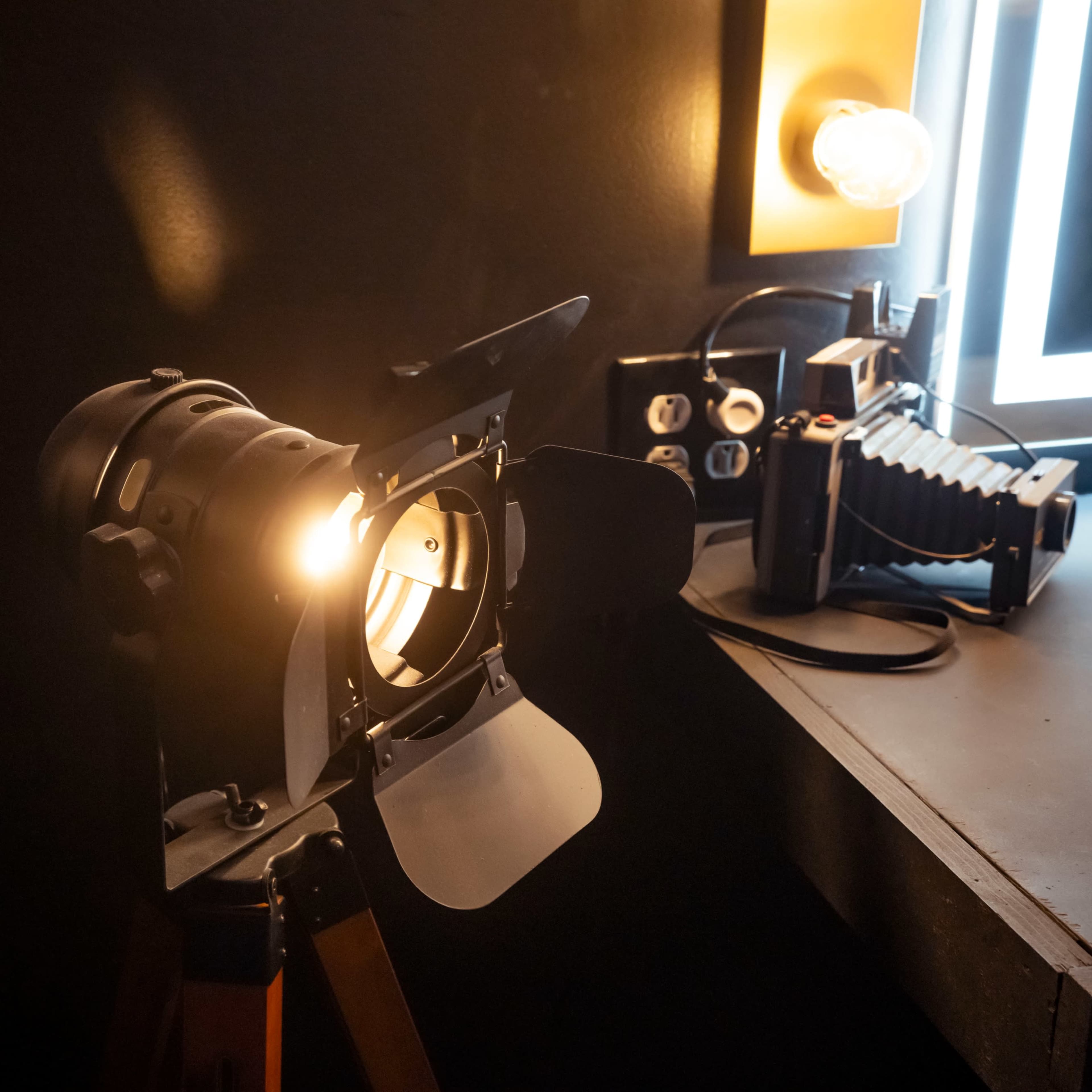 A studio light is positioned on a tripod next to a dark wall, with a camera and a vintage flash on the counter nearby.
