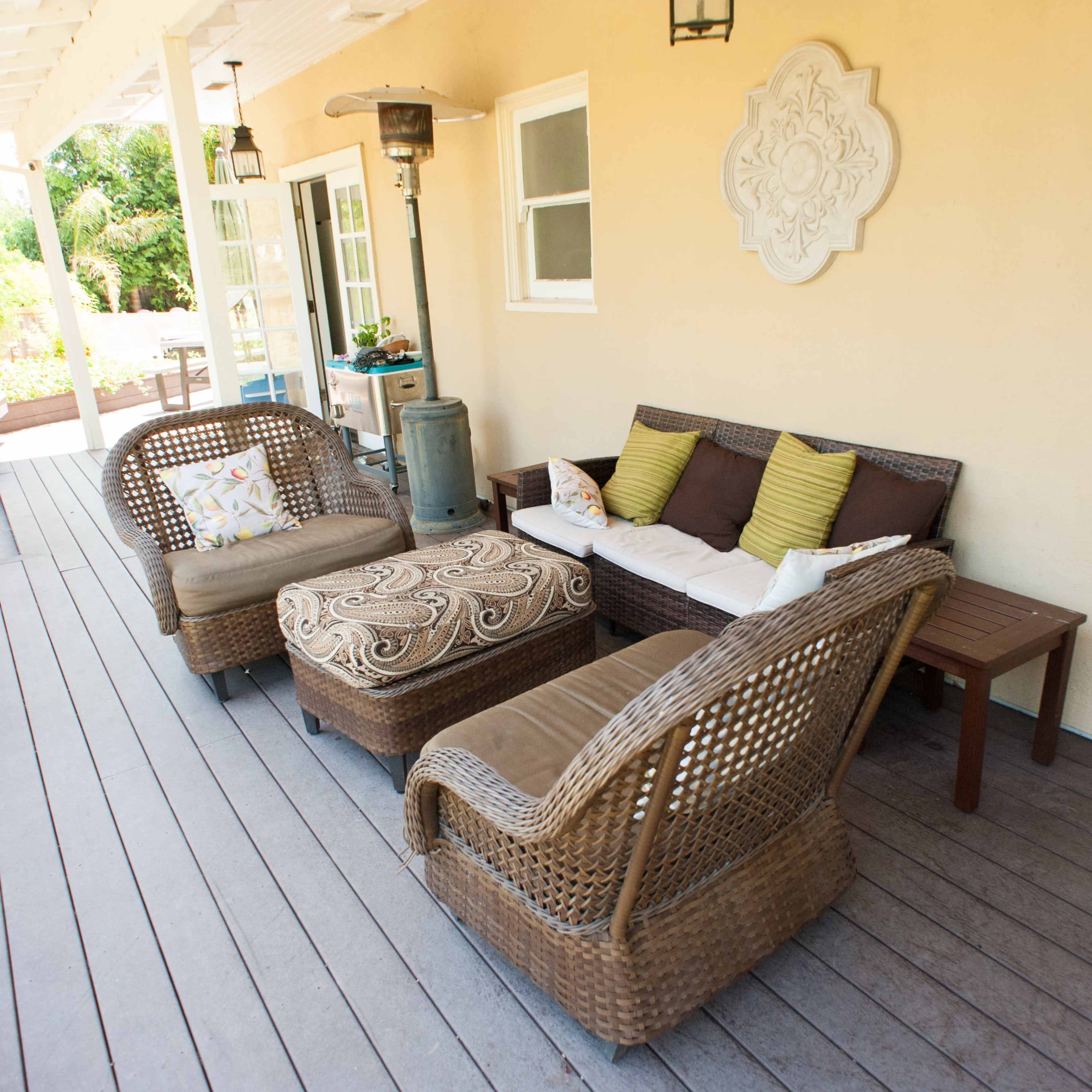 A covered outdoor seating area with wicker furniture, including two chairs, an ottoman, and a small table against a wall.