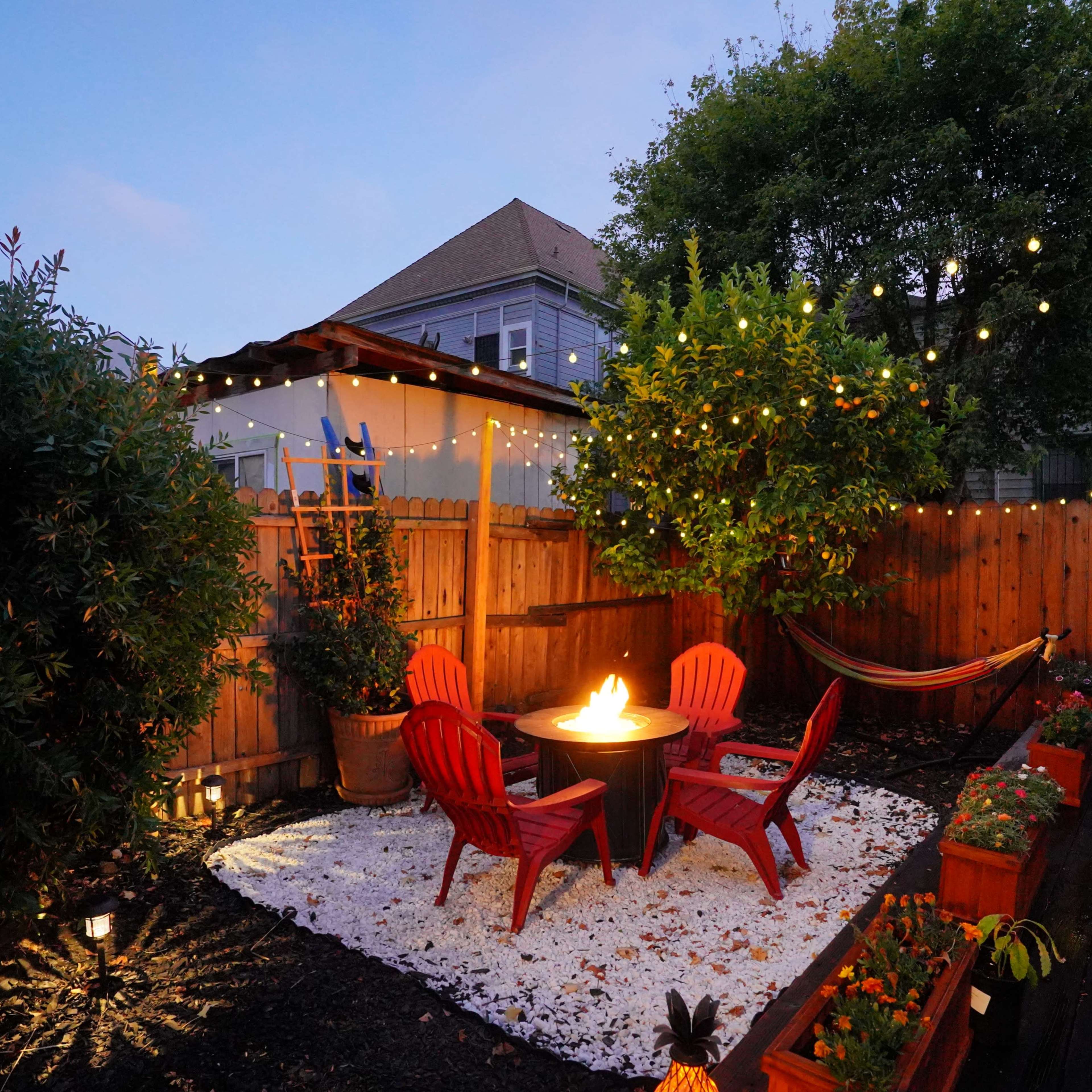 A small outdoor patio features red chairs around a fire pit, with potted plants along the edges and string lights overhead.