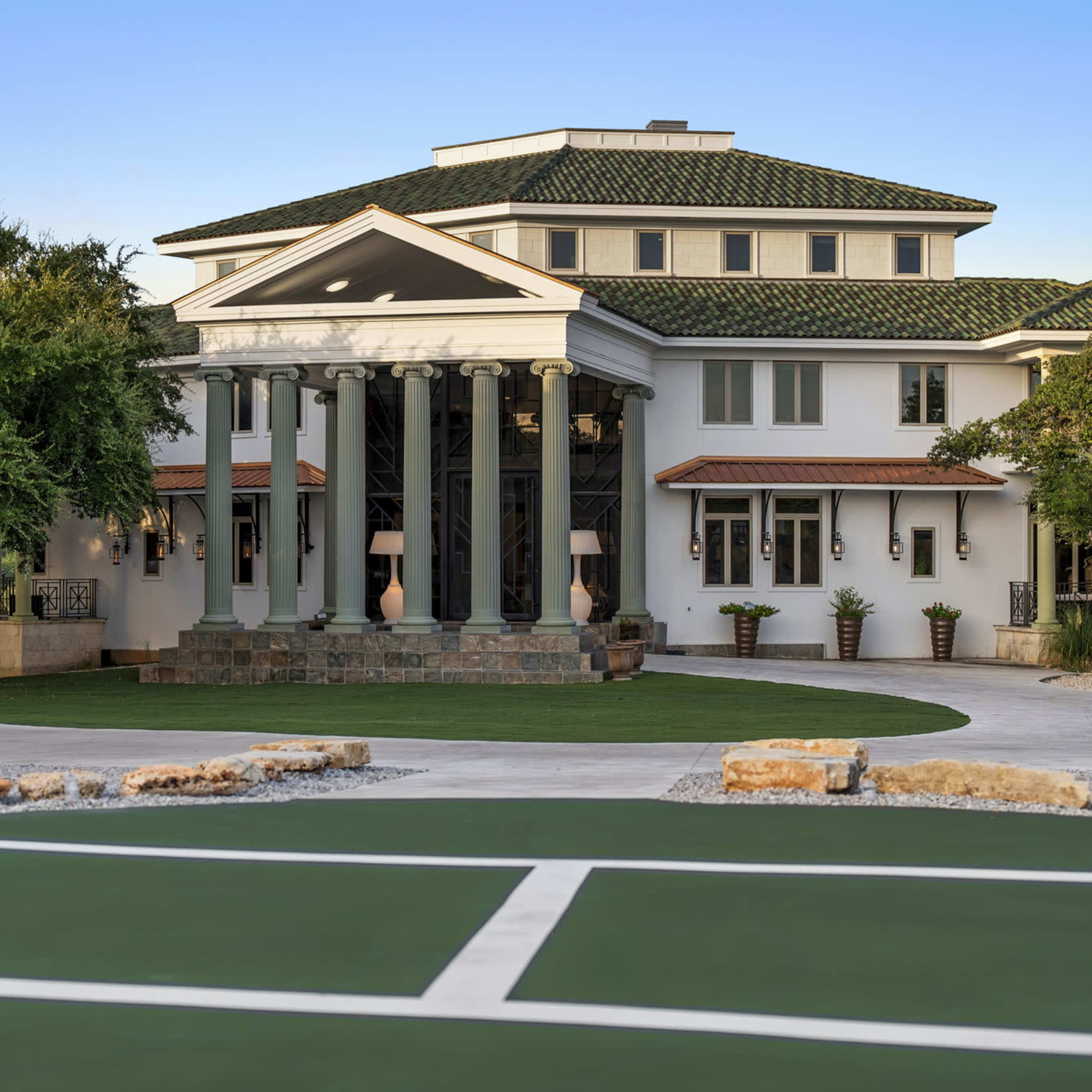 A large, modern mansion featuring a series of columns at the entrance and manicured landscaping.