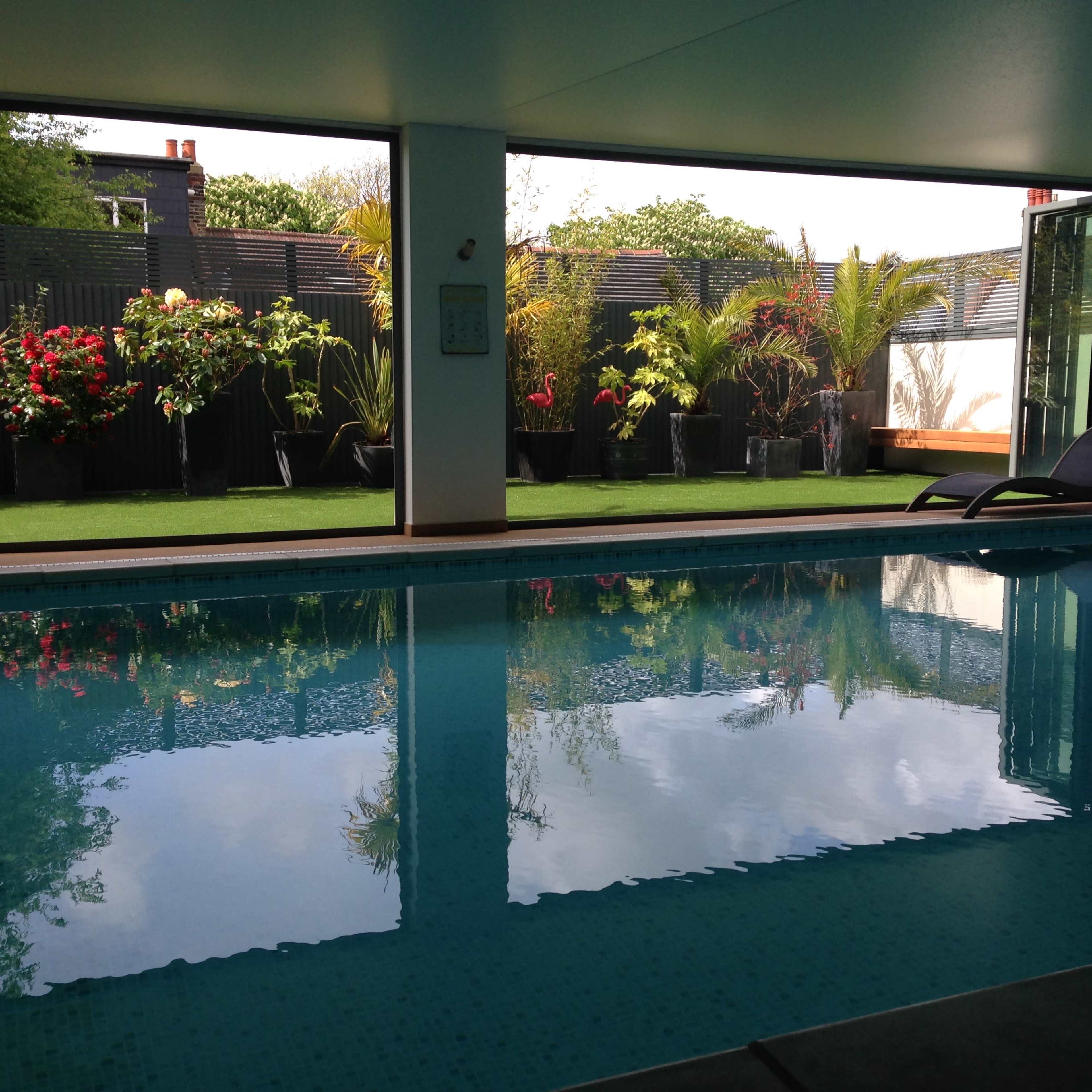 A swimming pool indoors, reflecting greenery and flower arrangements visible through large open windows.