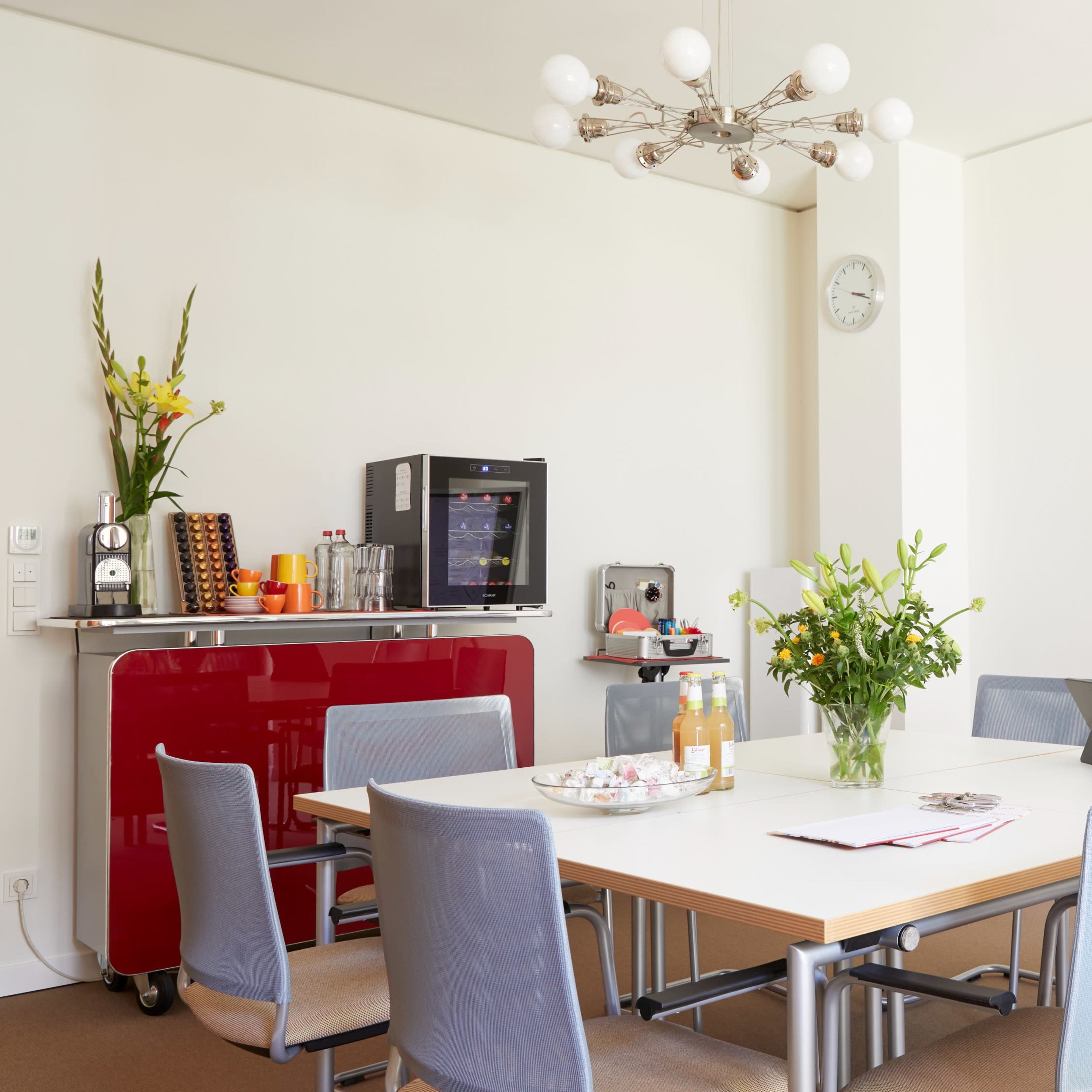 A modern meeting room features a large table surrounded by chairs, a display screen, and a small kitchenette with refreshments.