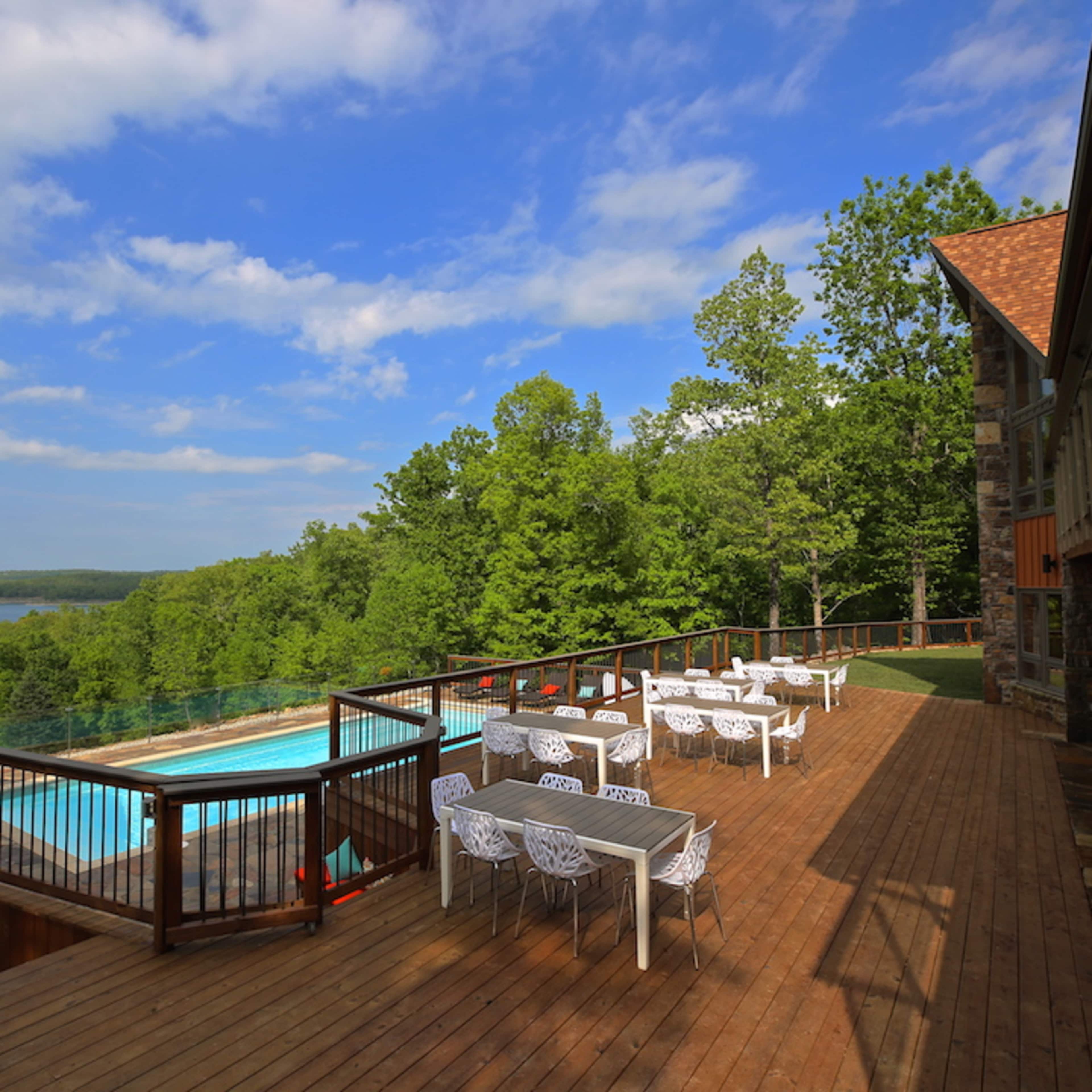A wooden deck with white tables and chairs overlooks a swimming pool and a scenic view of trees and a lake under a partly cloudy sky.