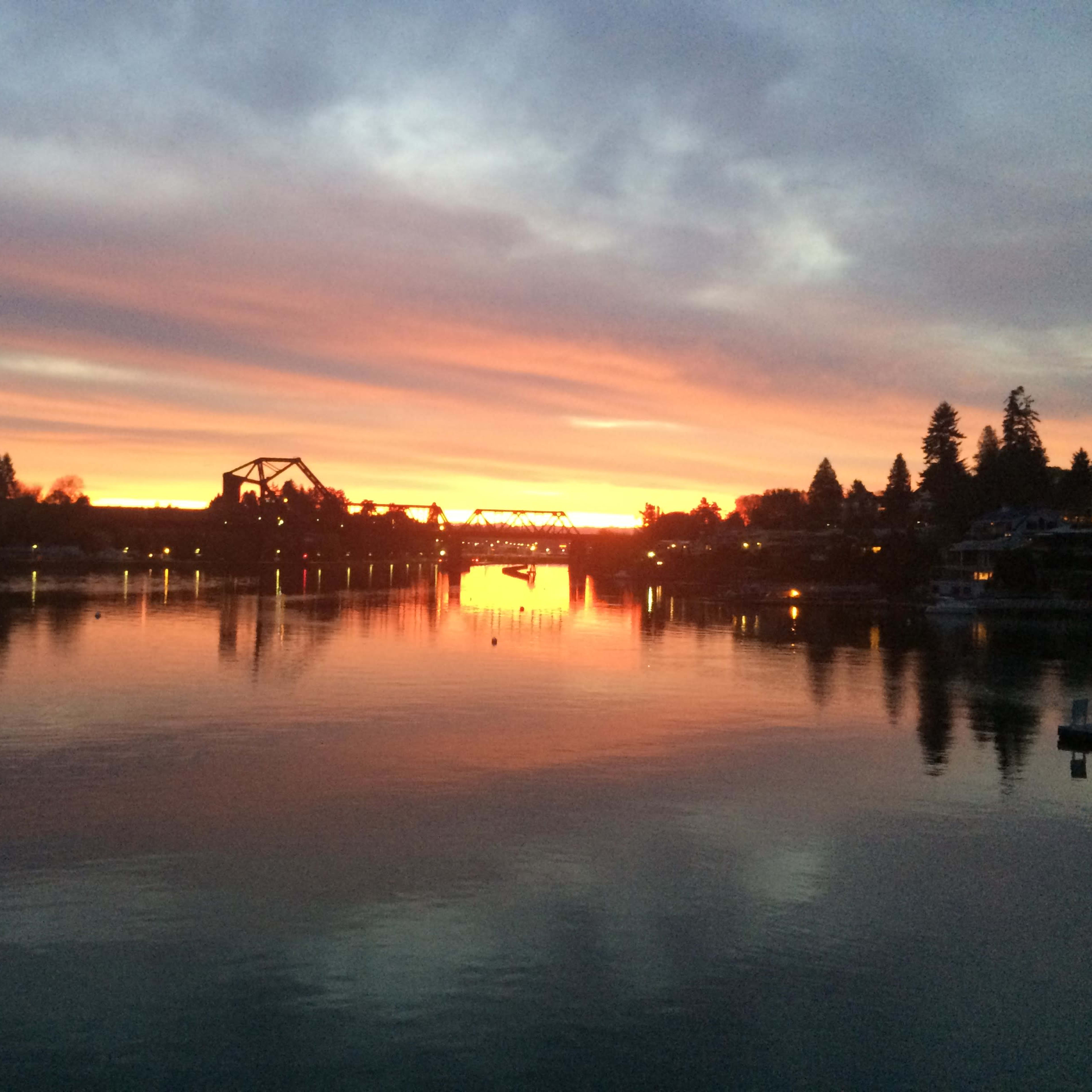 A river at sunset with a reflective surface and silhouettes of buildings and trees along the shore.