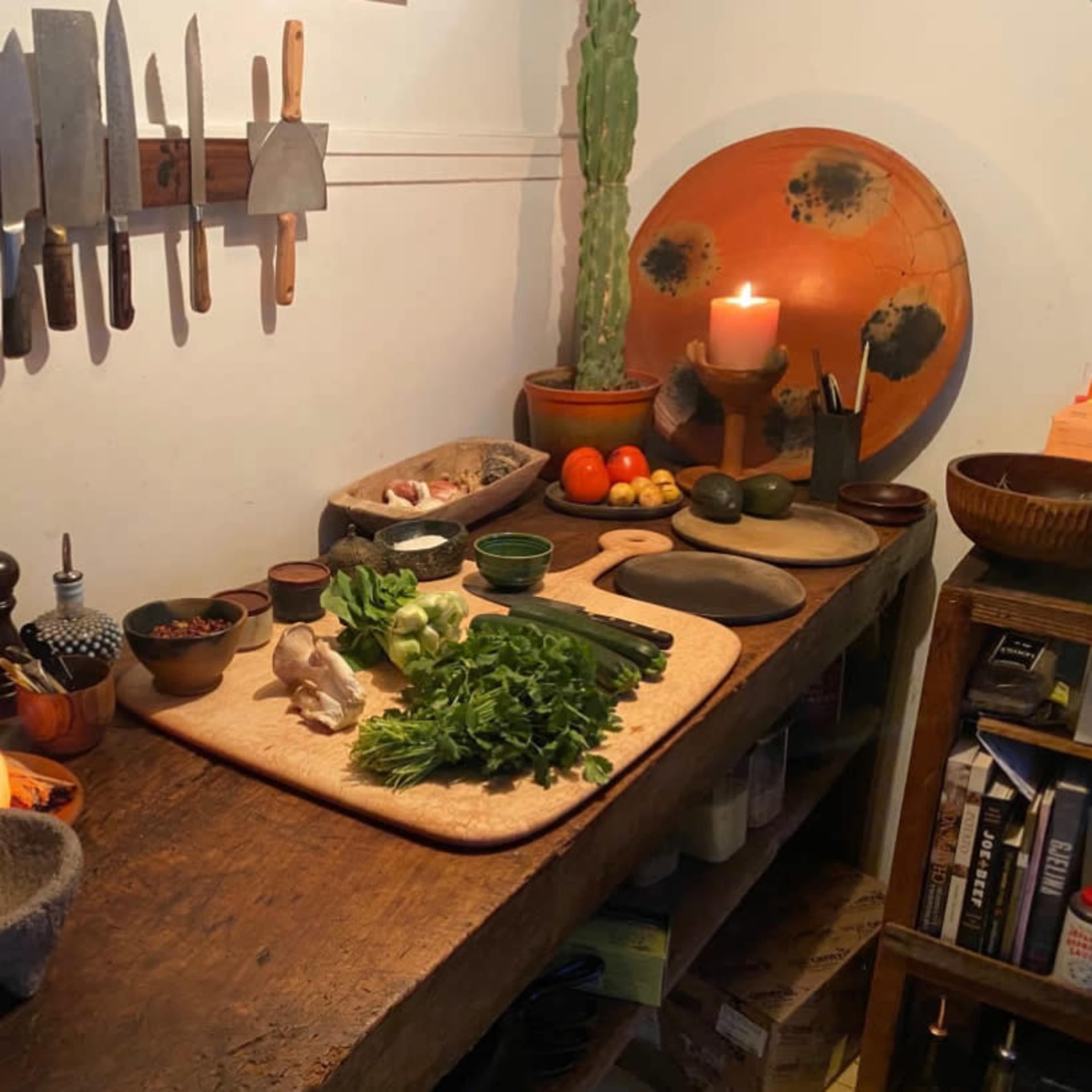 A wooden kitchen table is organized with various cooking utensils, fresh vegetables, spices, and a lit candle.
